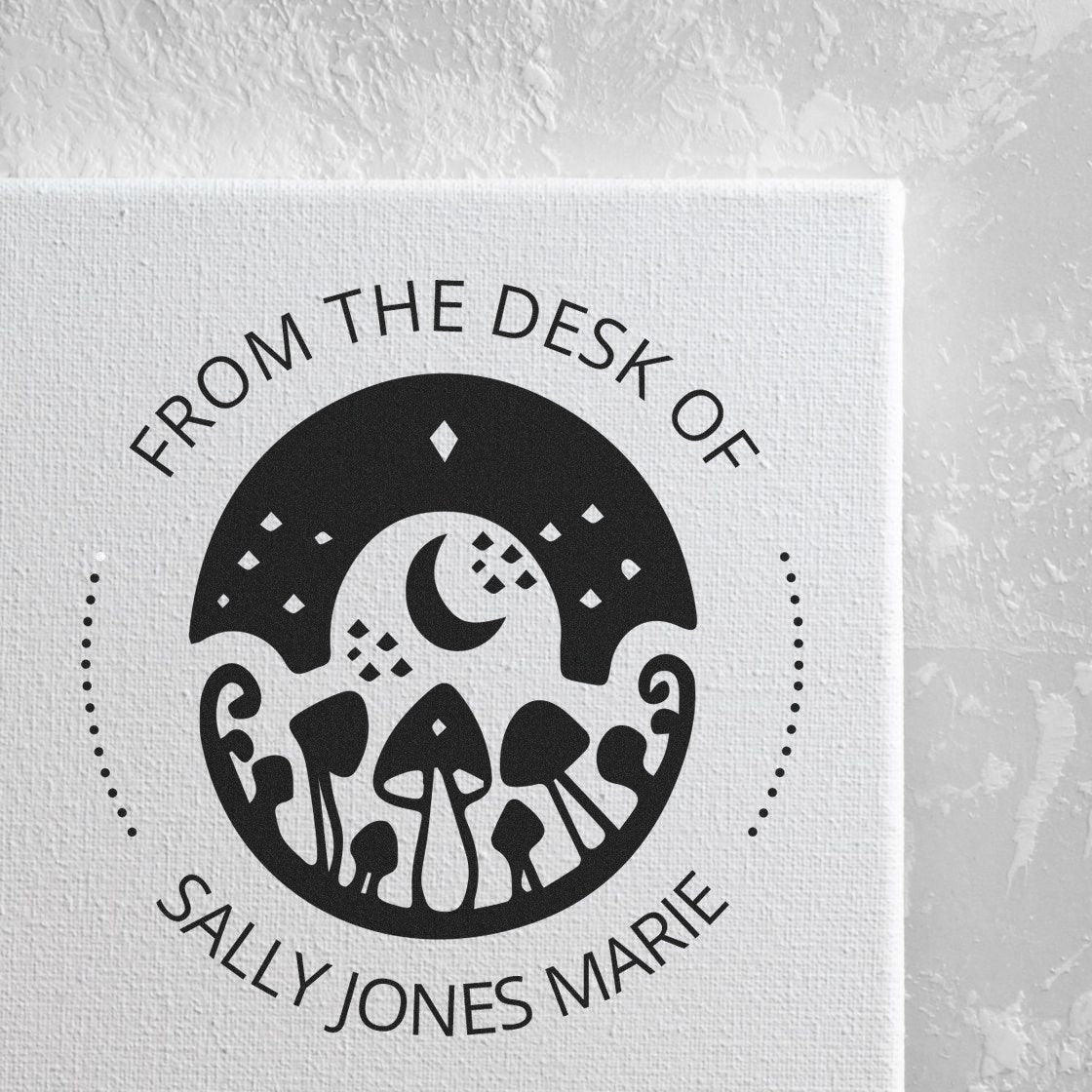 Black circular stamp seal with moon and mushrooms, reading From the Desk of Sally Jones Marie on white textured paper.