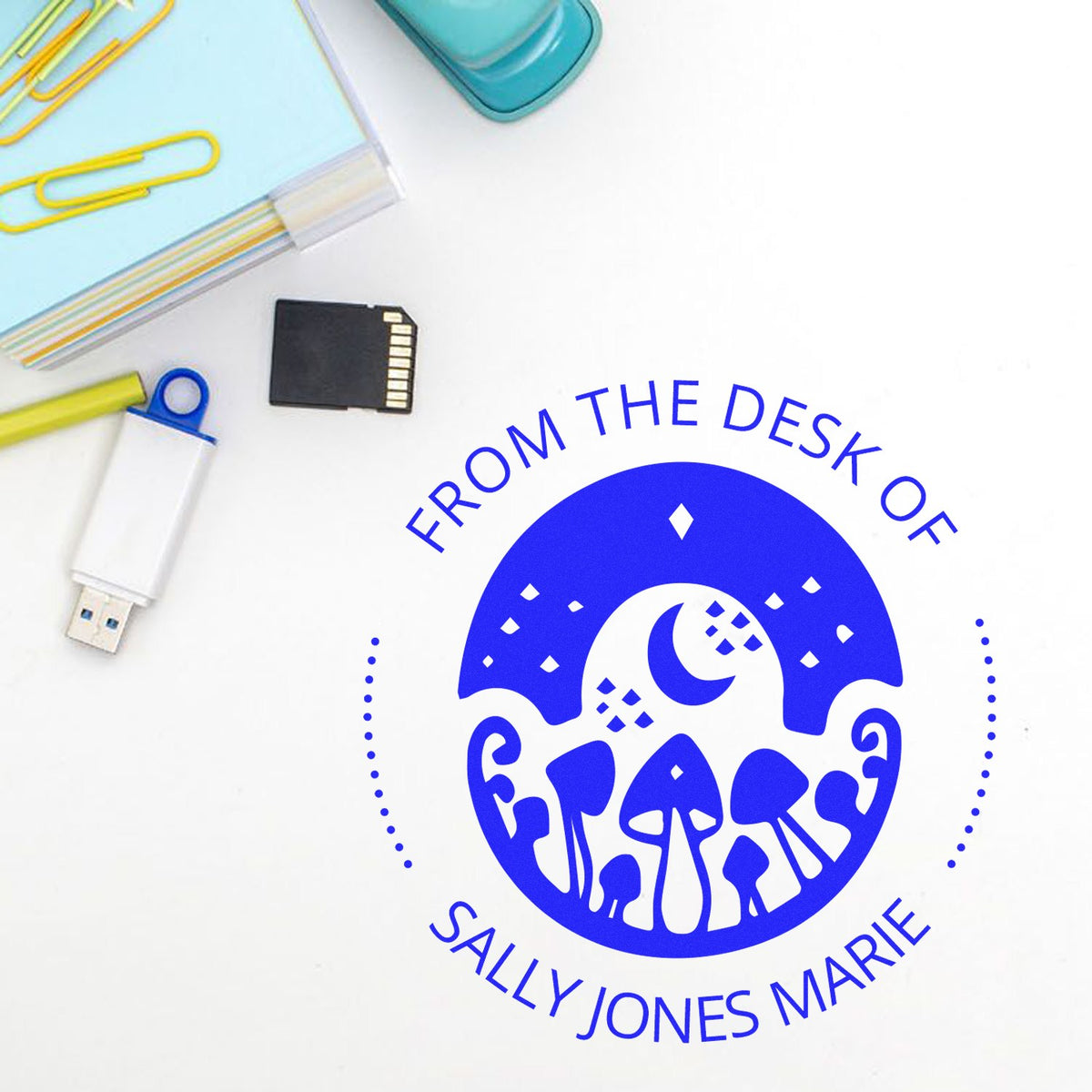 Blue custom desk stamp seal with mushroom moon design, beside paper clips, USB drive and SD card.