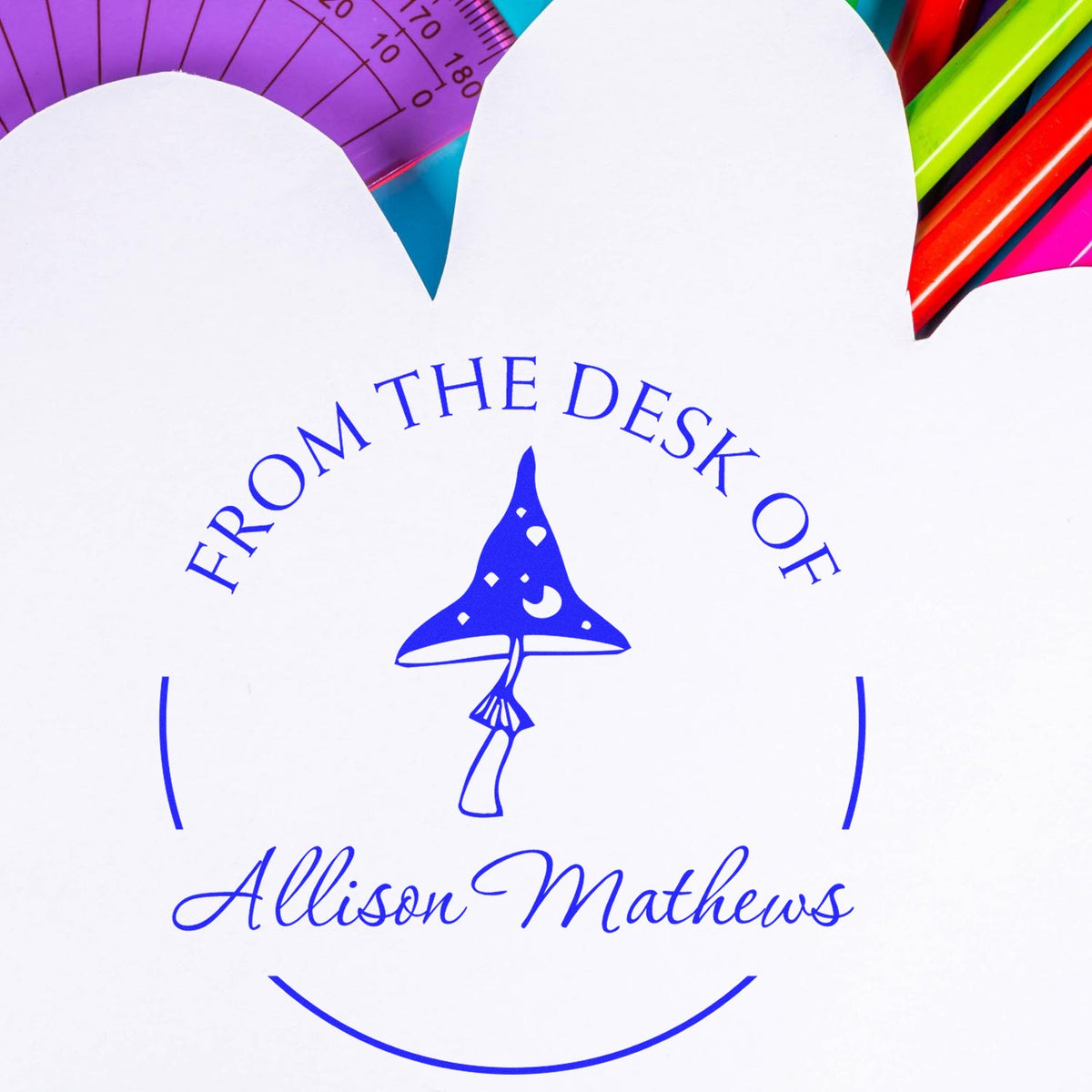 White custom desk stamp/seal with blue From the Desk of Allison Mathews text, mushroom icon, and colorful pens.