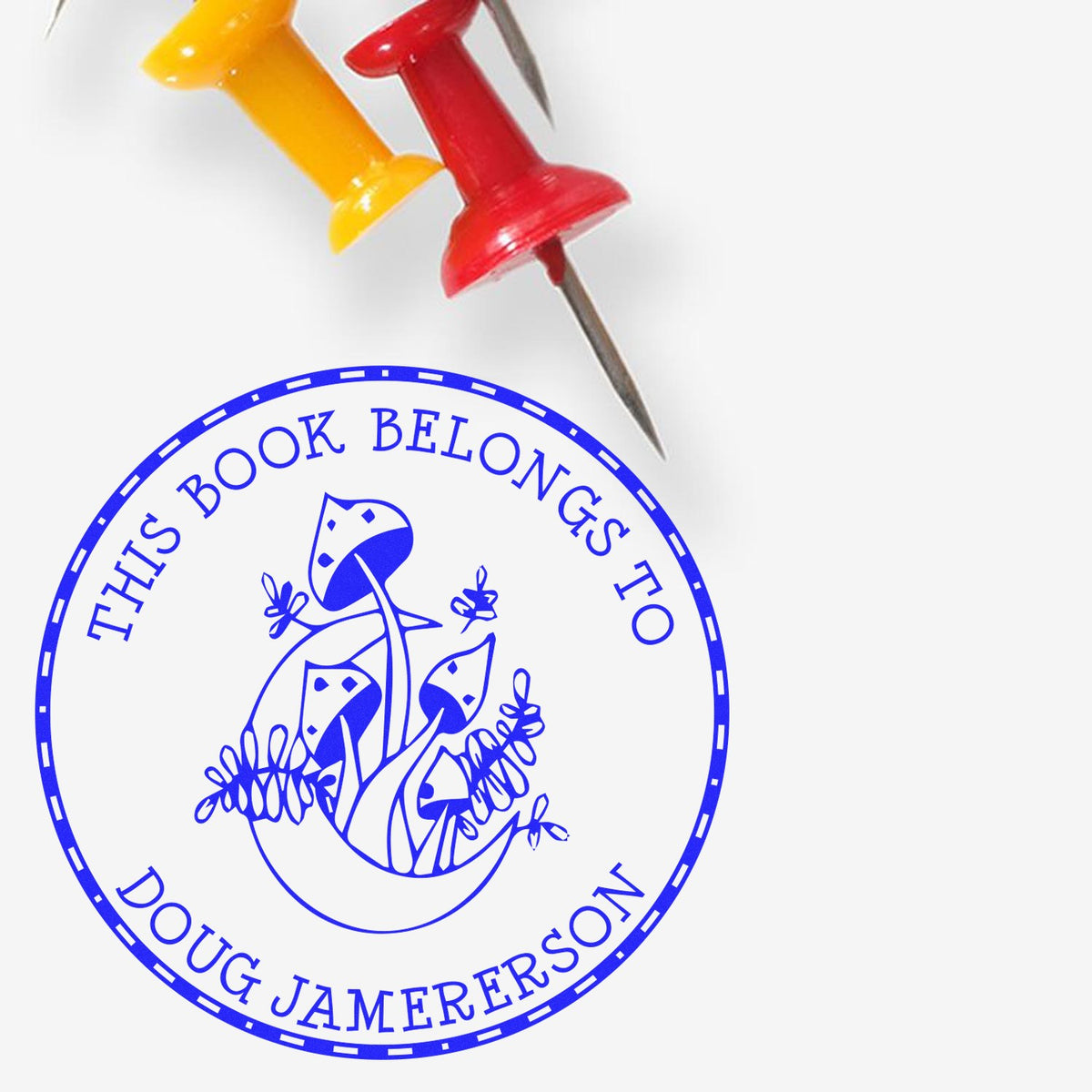 Blue personalized book stamp seal with mushrooms, reading ÂThis Book Belongs to Doug Jamerson, beside red and yellow pushpins.