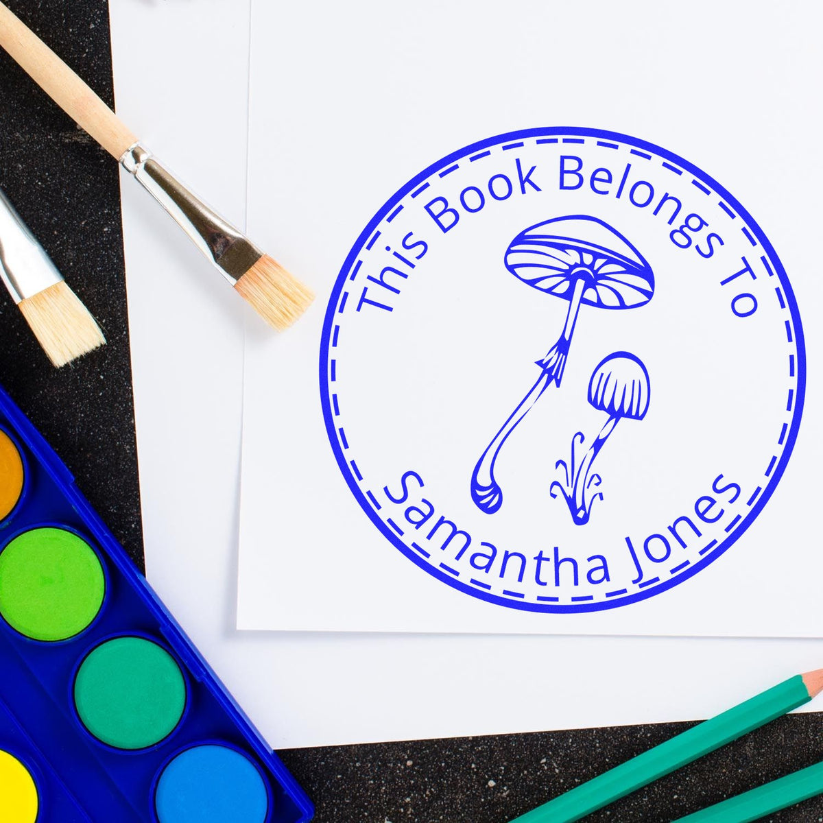 Blue custom book stamp seal with mushroom design and "This Book Belongs To Samantha Jones" beside paintbrushes and watercolor palette.