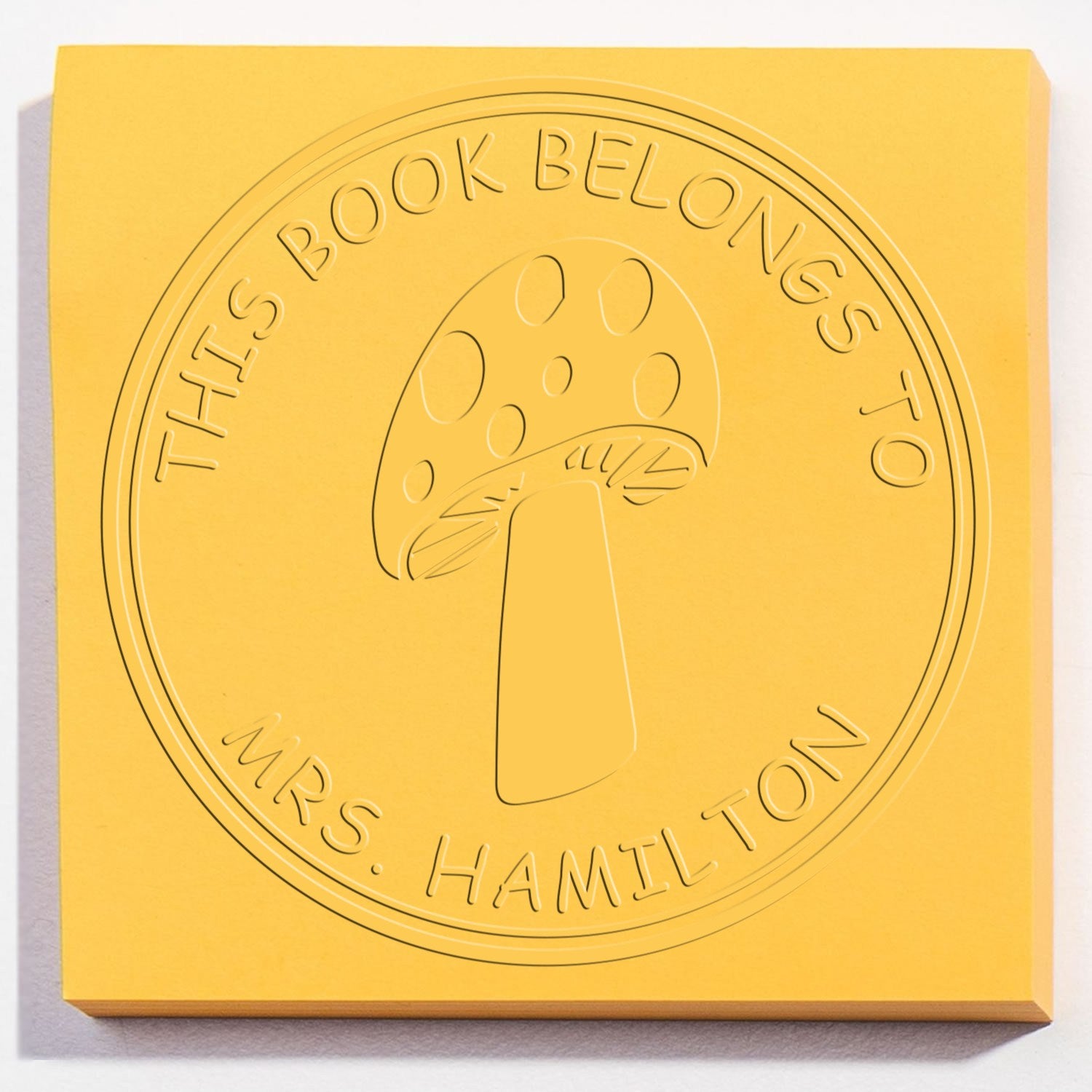 Yellow embossed book stamp seal with mushroom icon and This Book Belongs to Mrs. Hamilton text.