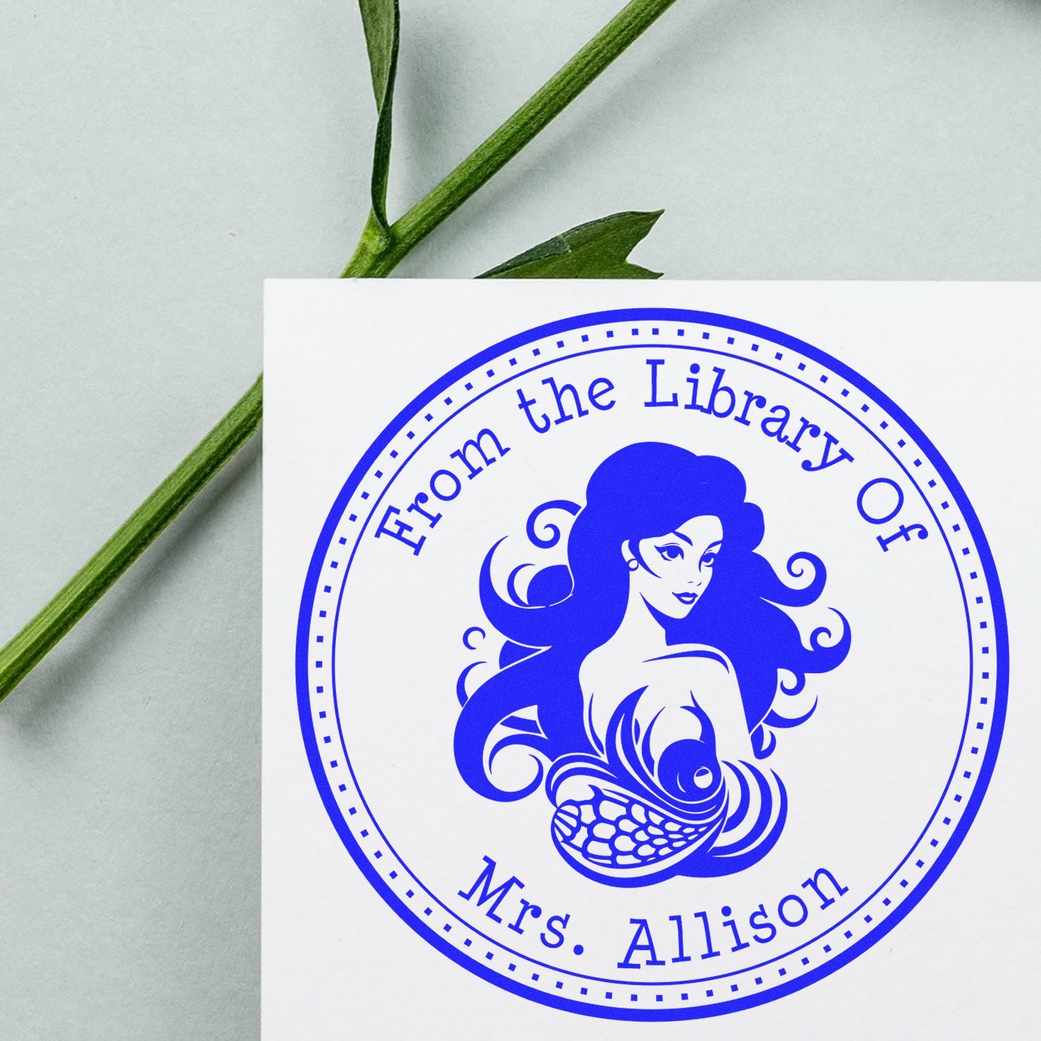 Blue custom library seal stamp with mermaid design and text From the Library Of Mrs. Allison on white card.