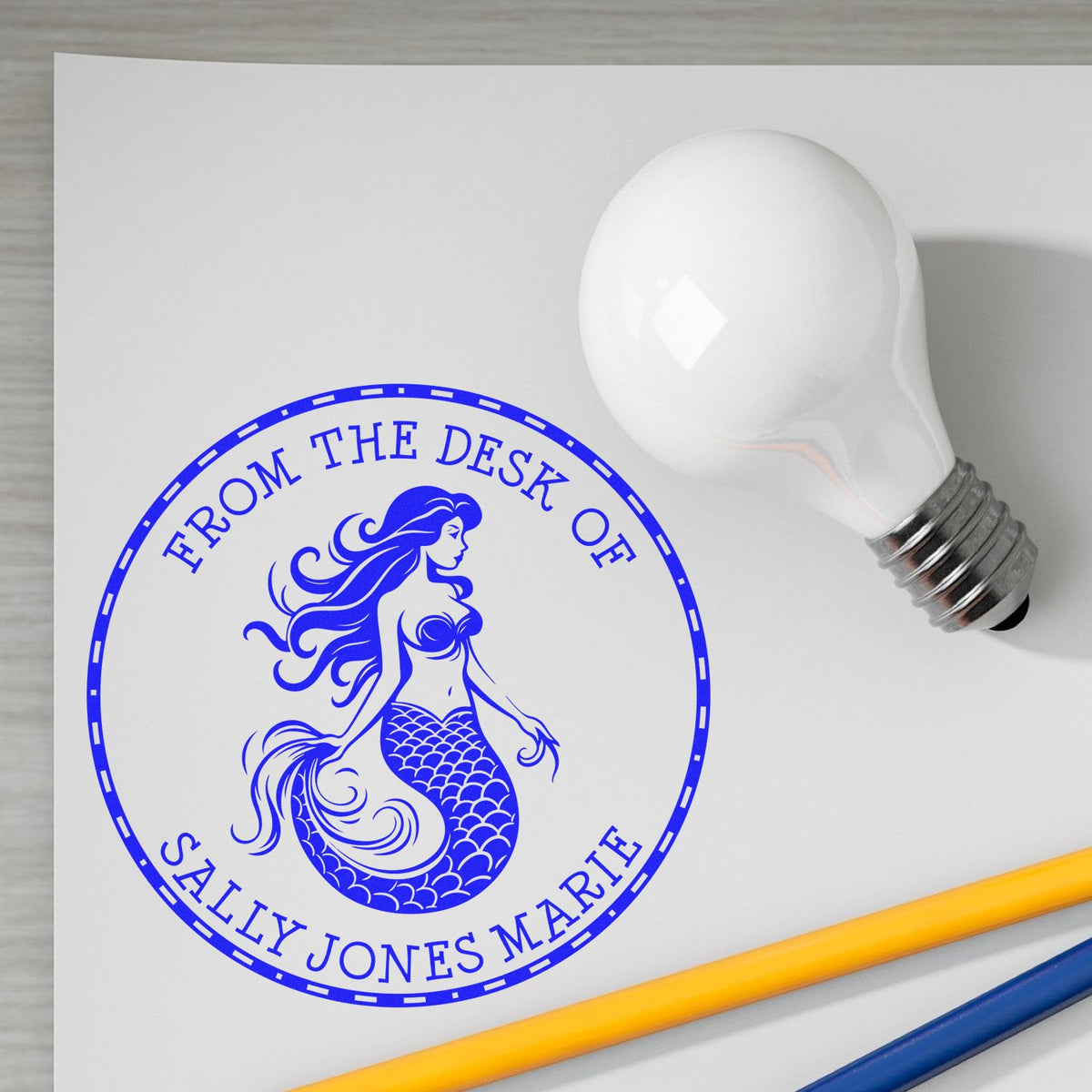 Blue personalized mermaid stamp seal reading From the desk of Sally Jones Marie on white paper, beside a light bulb and pens