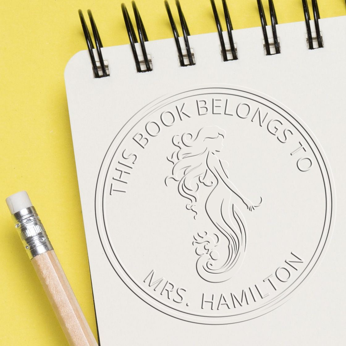 Embossed white personalized bookplate stamp seal with mermaid design on yellow background, beside a pencil.