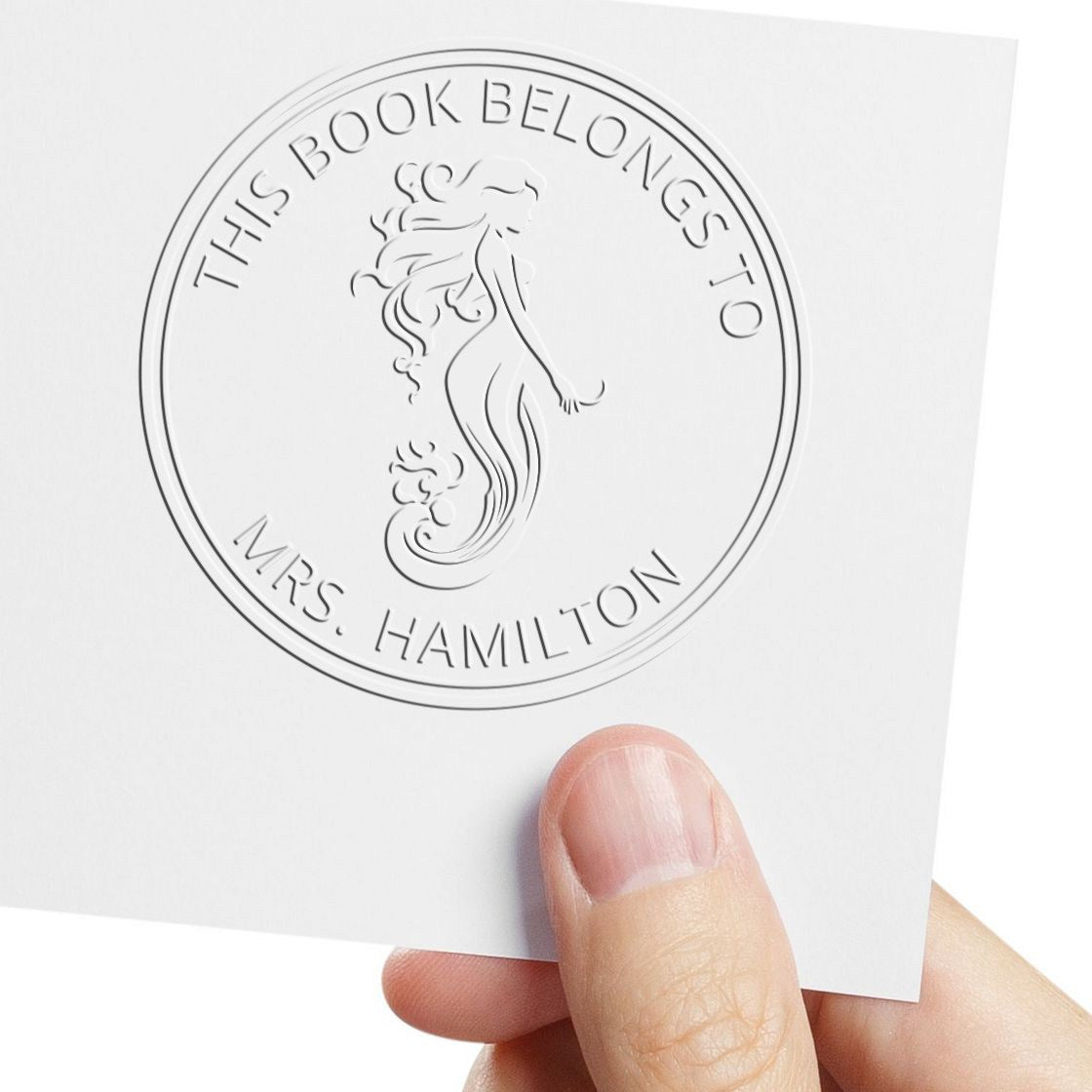 White embossed circular stamp seal with mermaid, reading This Book Belongs to Mrs. Hamilton, held on a white card.