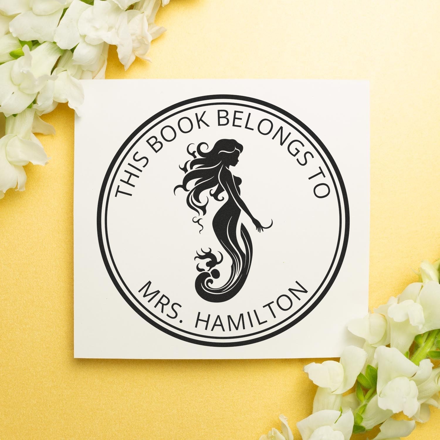 Black round mermaid stamp seal reading This Book Belongs to Mrs. Hamilton on white card, with pale yellow background and white flowers.