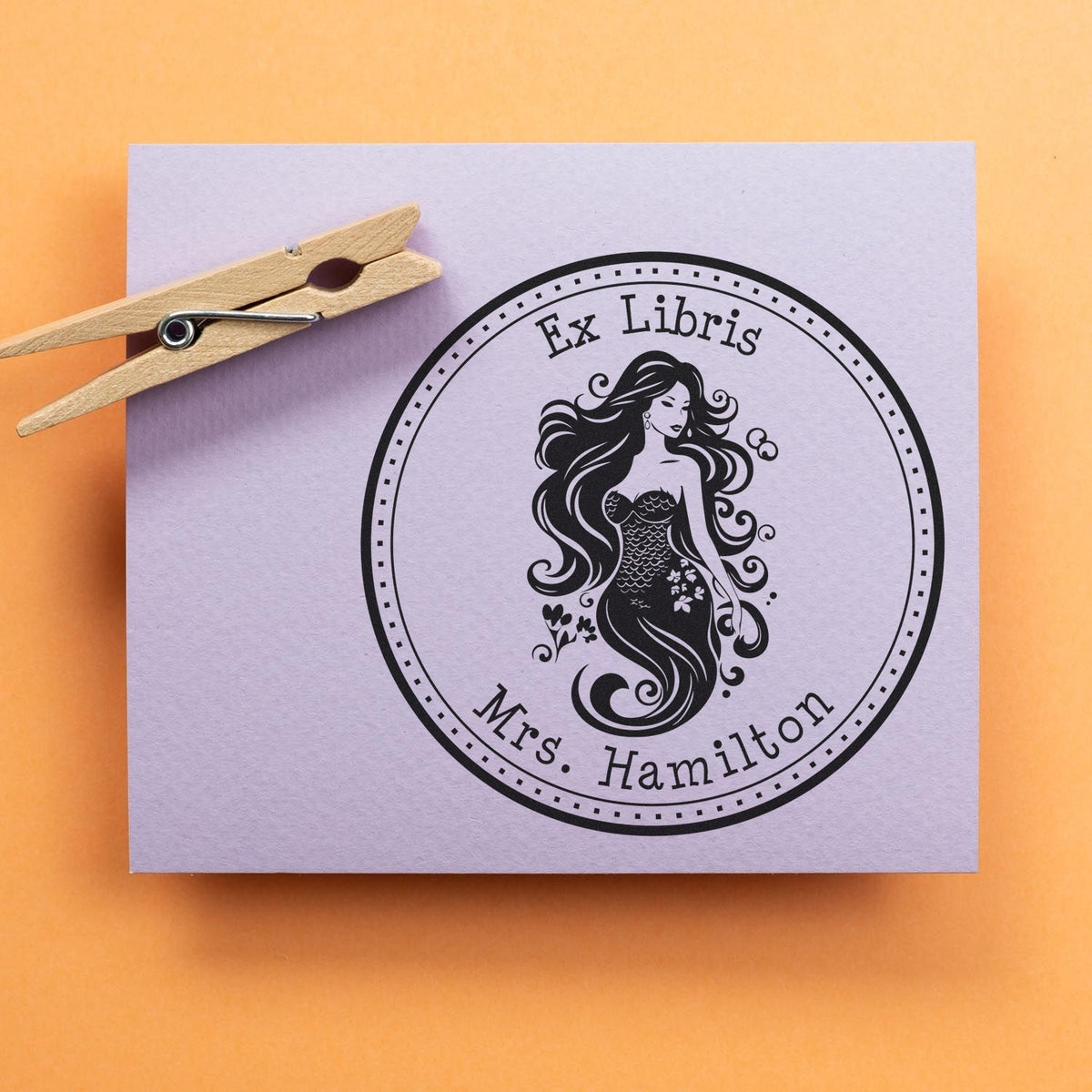 Wooden clothespin beside a lavender ex libris stamp seal with a black mermaid and Mrs. Hamilton on orange background.