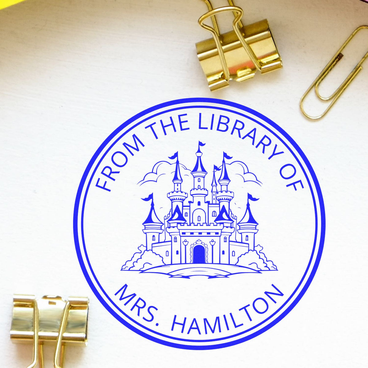 Blue circular library stamp seal with castle design and From the Library of Mrs. Hamilton, on white paper with gold binder clips.