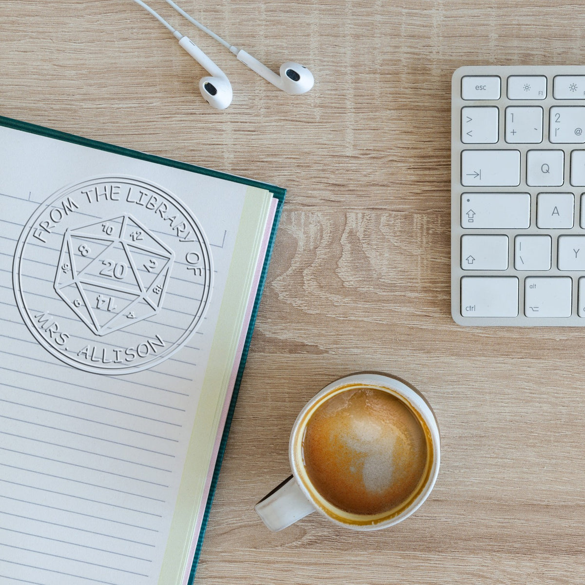 Custom embossed library stamp seal on a lined notebook beside a white keyboard, earbuds, and coffee on a wood desk.