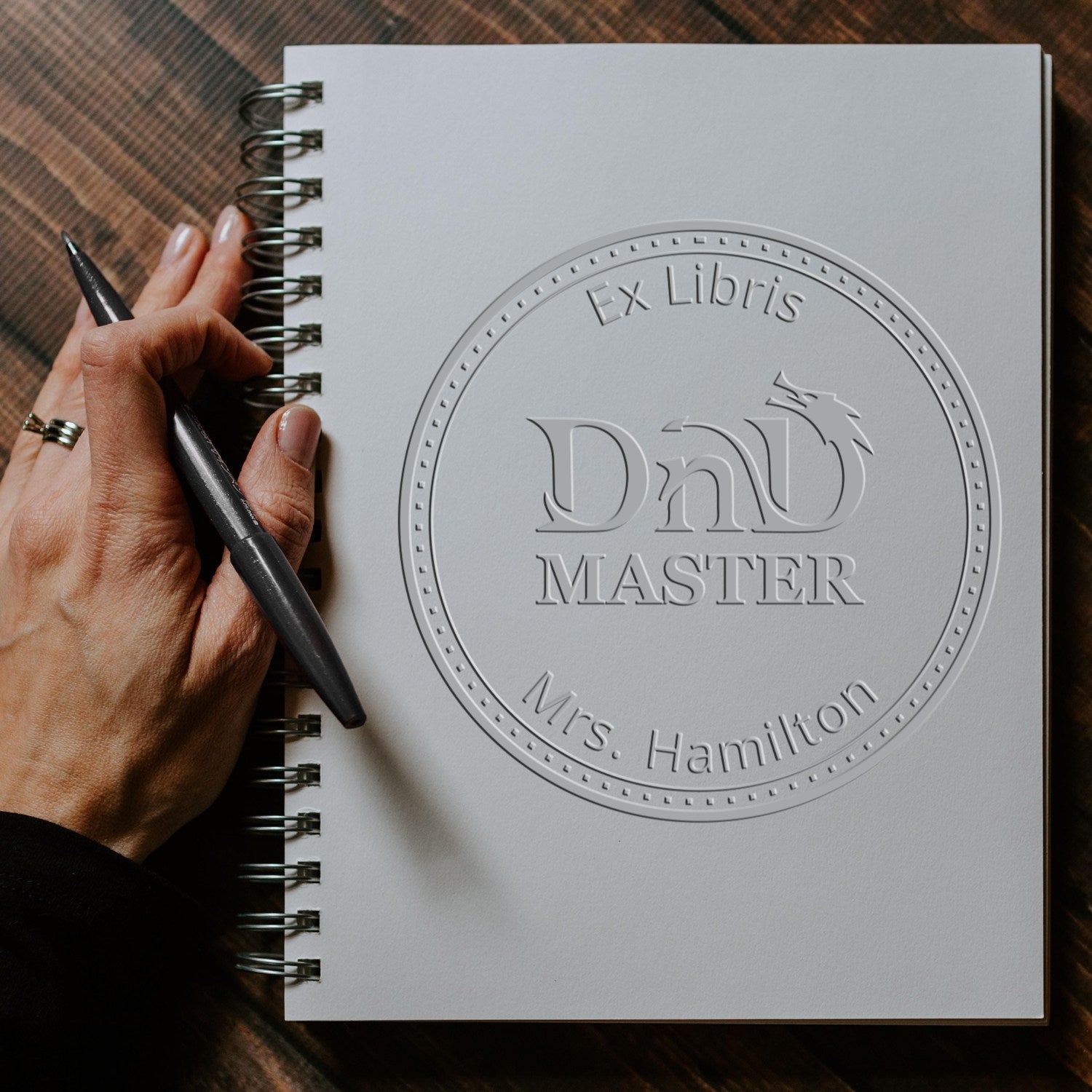 White spiral notebook featuring a silver embossed custom seal stamp, held by a hand with a black pen.