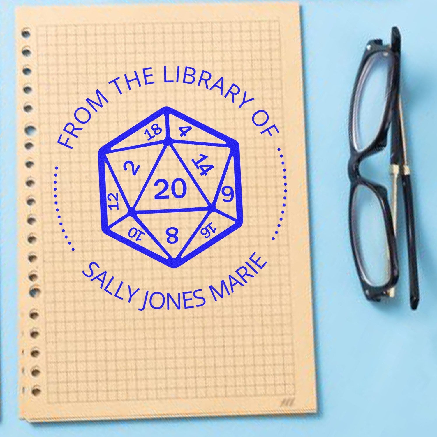 Blue custom library stamp seal with geometric design on beige graph paper notebook, beside black eyeglasses.