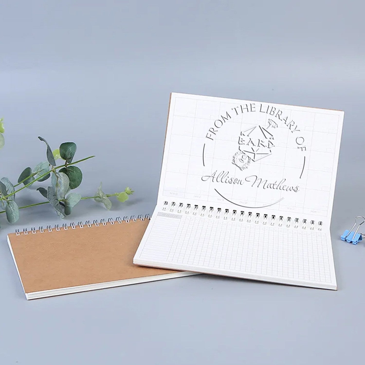 White custom stamp and seal notebook with embossed library logo, paired with brown cover and eucalyptus on a gray background.