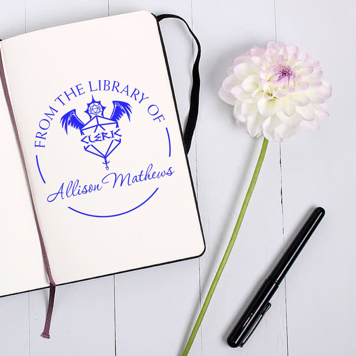 Blue custom From the Library of Allison Mathews stamp on a cream notebook, with a white flower and black pen.