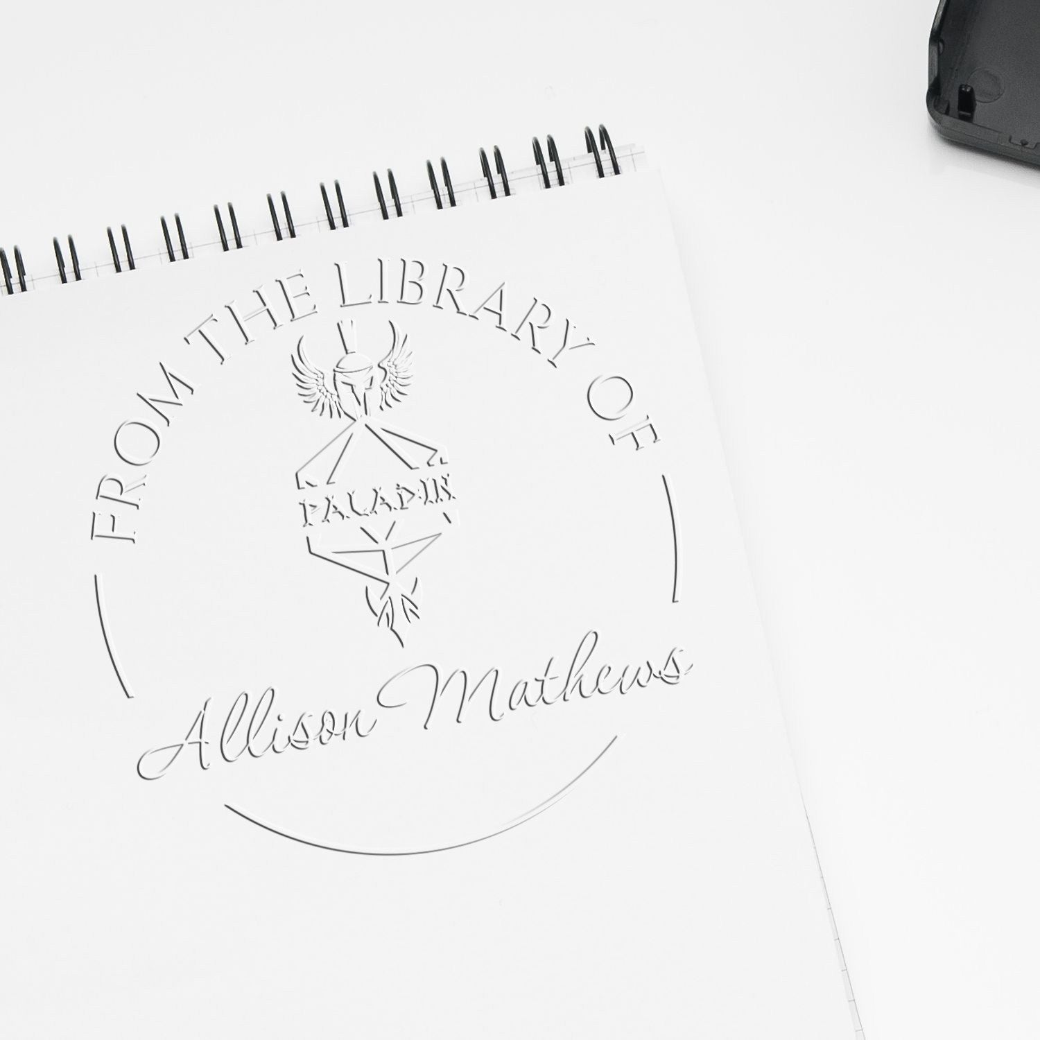 Embossed white library seal stamp on spiral notebook, reading From the Library of Allison Mathews, with black stamp device.