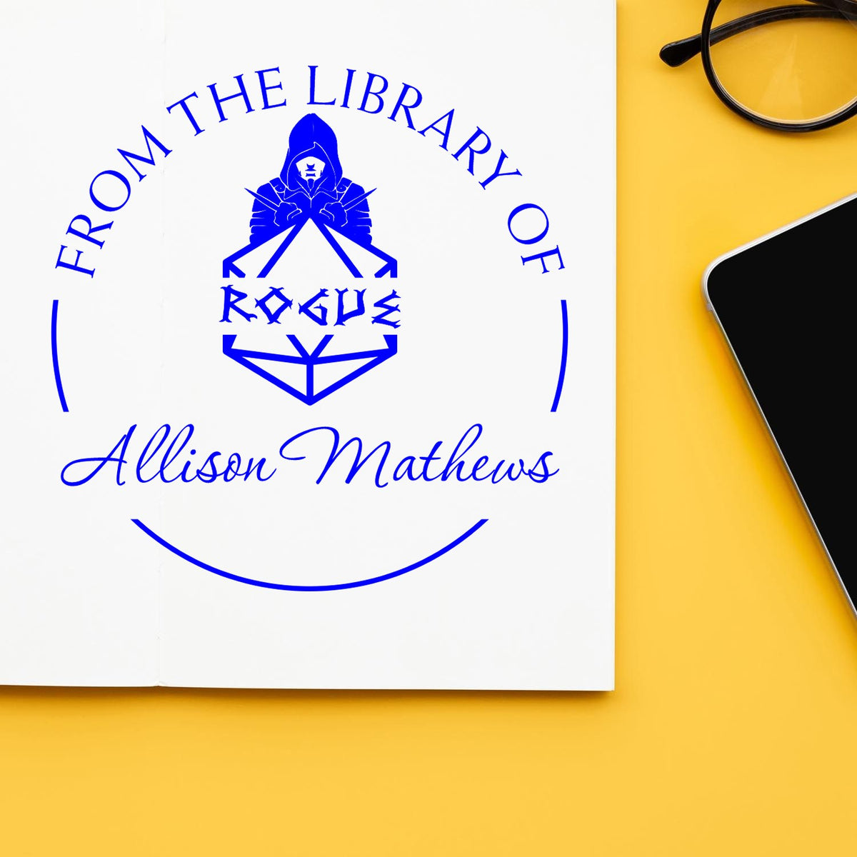 Blue custom library rubber stamp seal with hooded emblem and Allison Mathews name on white paper, beside glasses on yellow background.