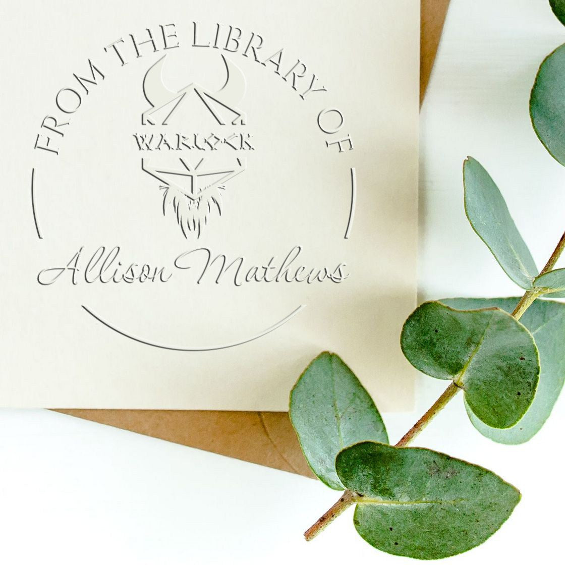 Cream custom library stamp seal reading From the Library of Allison Mathews, with embossed black text and green eucalyptus leaves.