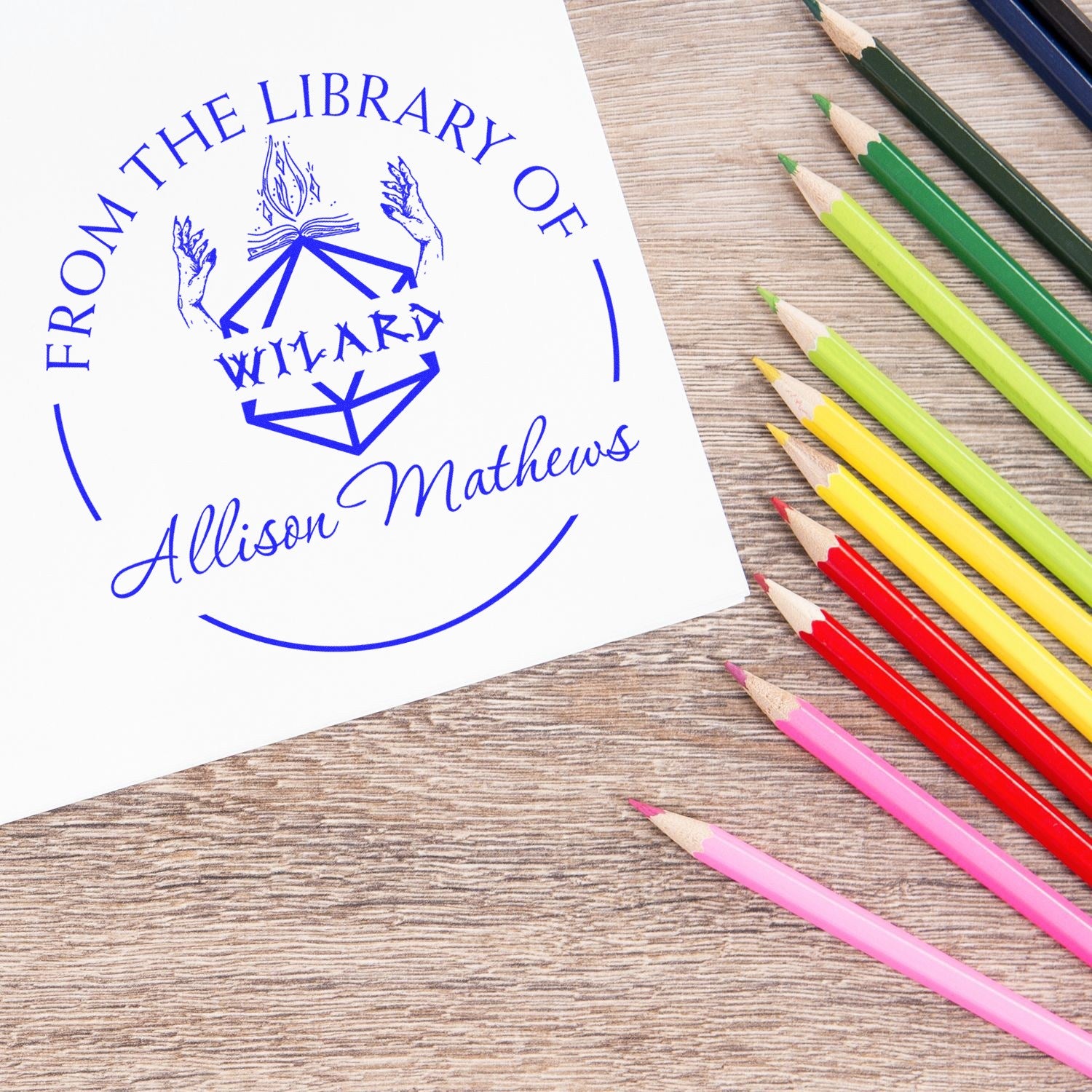Blue custom From the Library of wizard stamp seal on white paper beside rainbow colored pencils.