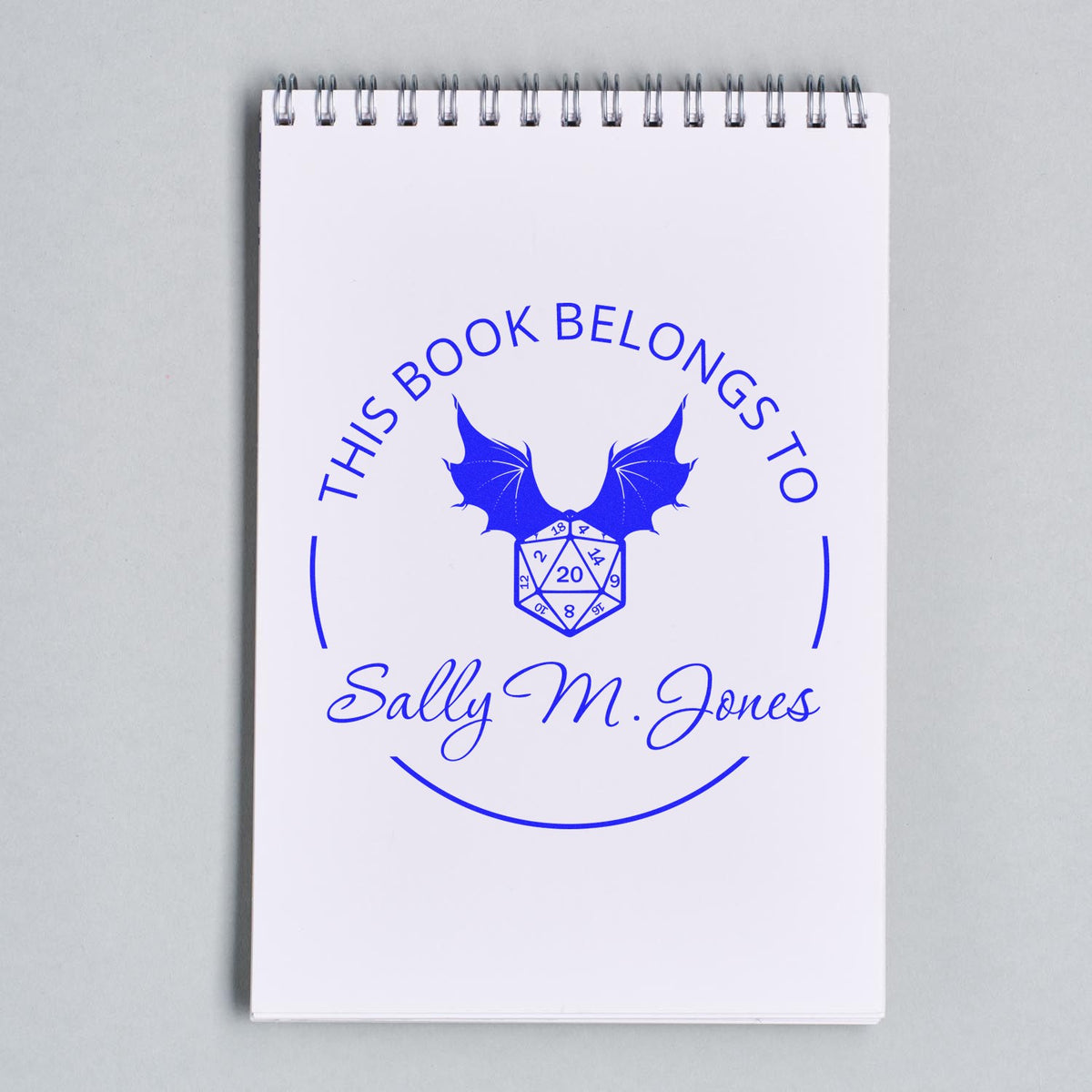 Blue custom stamp and seal on white spiral notebook, featuring bat wings, dice, and Sally M. Jones.