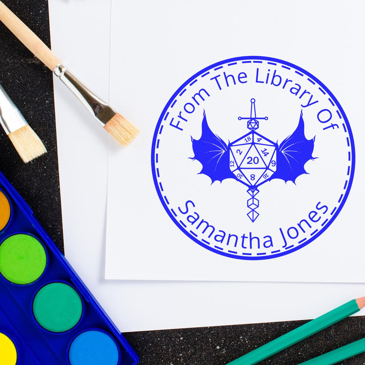 Blue custom stamp seal reading "From The Library Of Samantha Jones" with bat and d20, beside paint brushes and watercolor palette.
