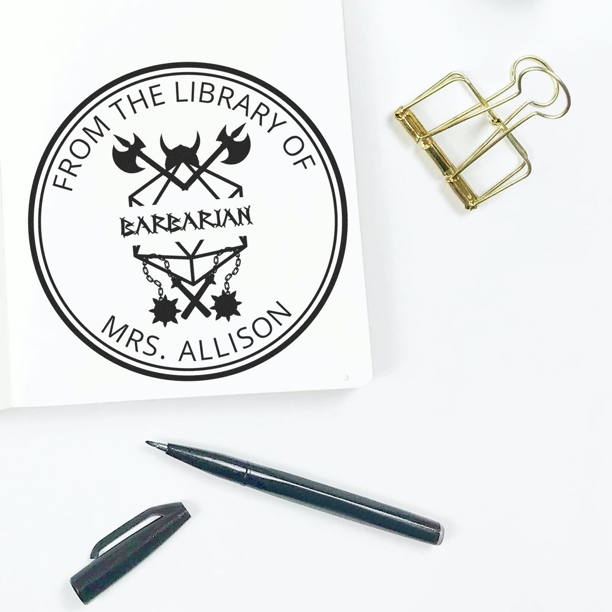 Custom black library stamp seal on white paper, with gold binder clip and black pen, reading ĀFrom the Library of Barbarian Mrs. AllisonĀ