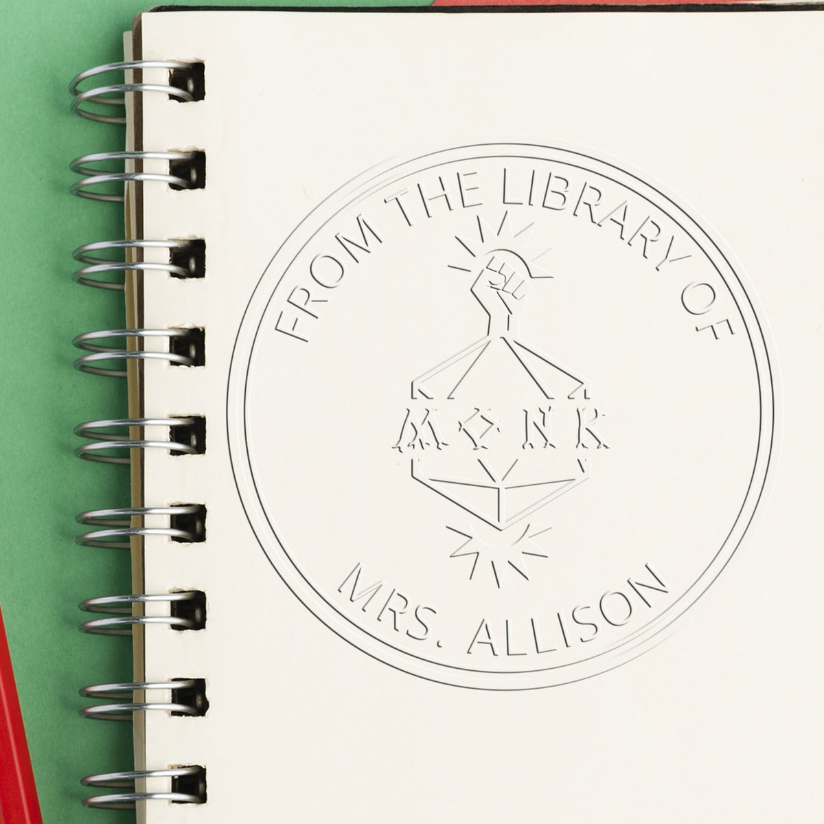 Custom embossed library seal stamp on cream spiral-bound notebook, silver rings on green background.