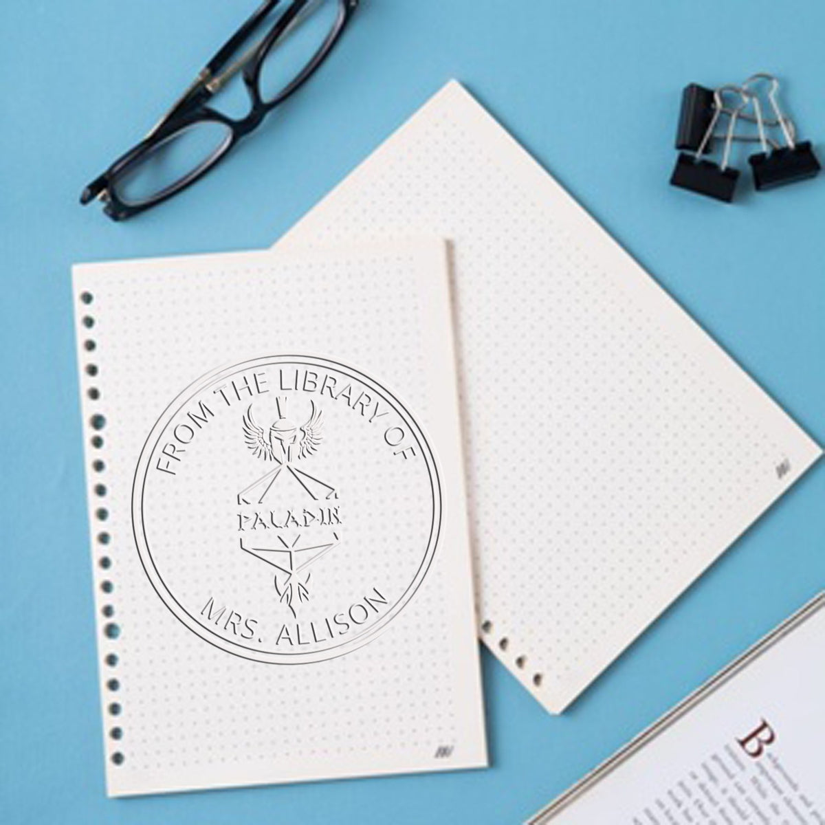 White dotted notebooks on blue with a custom circular library stamp seal, plus black glasses and binder clips.