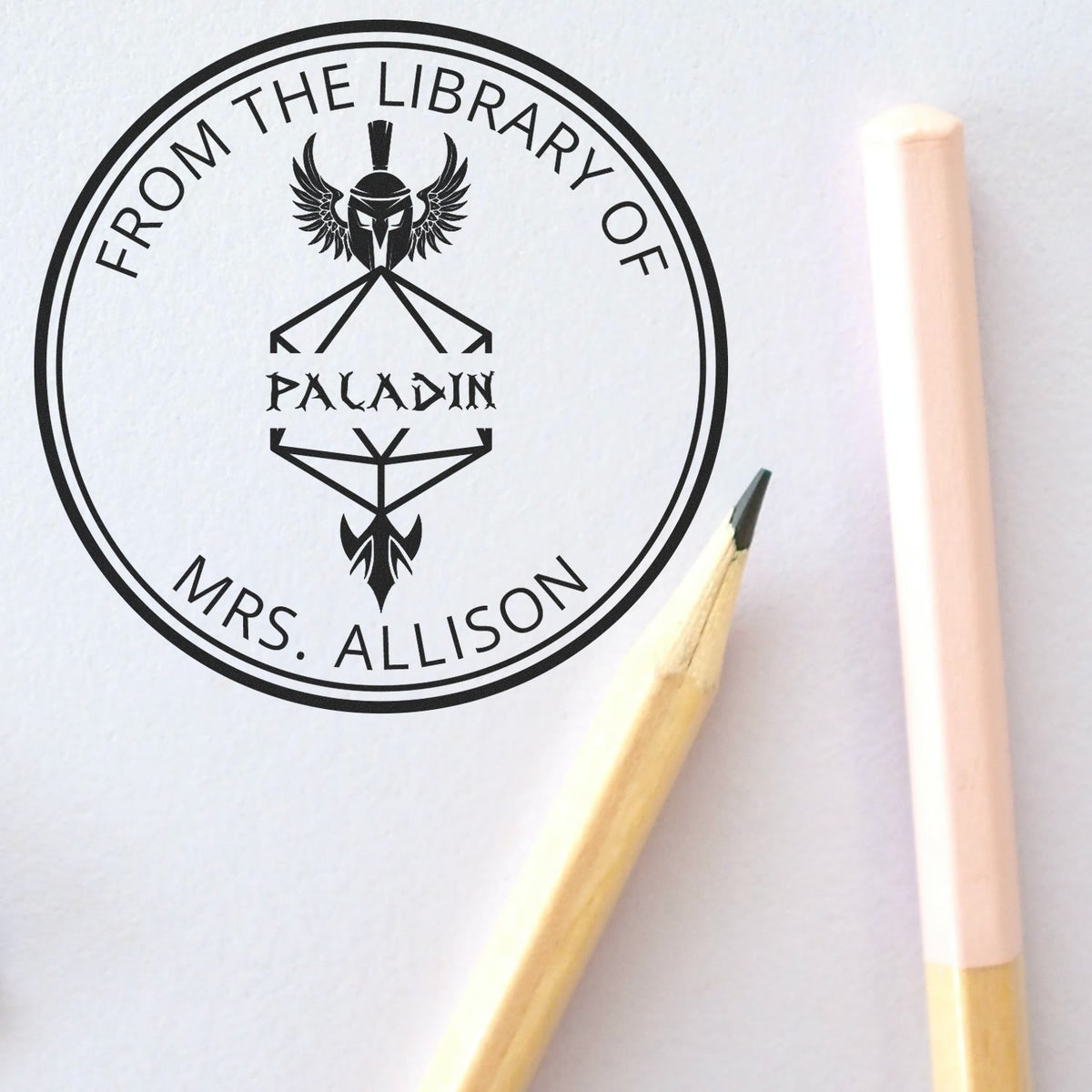 Black custom library seal stamp for Mrs. Allison, with Paladin emblem, on white paper beside pale pink and wood pencils.