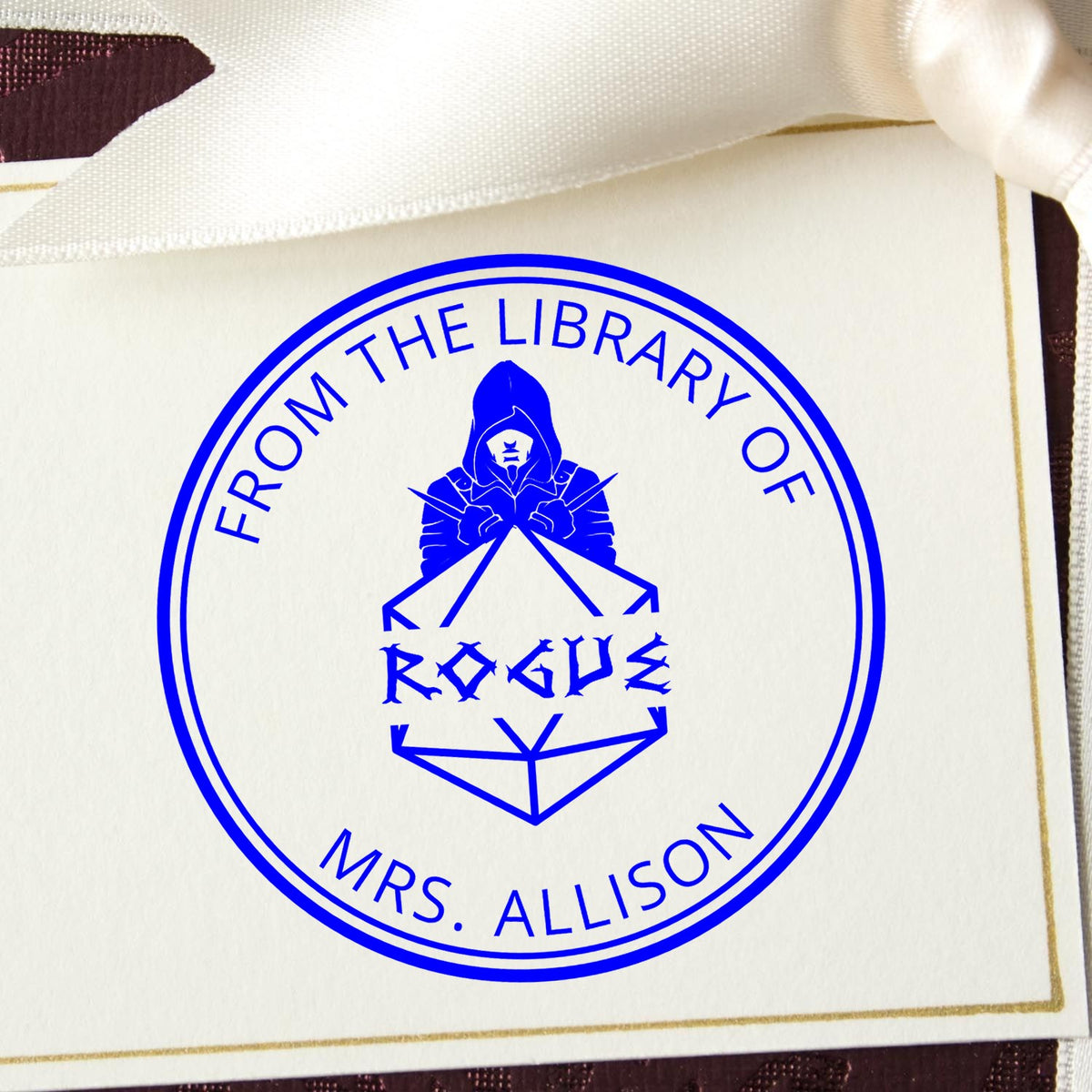 Blue circular library stamp seal with hooded figure and Rogue text on cream card, tied with white ribbon.