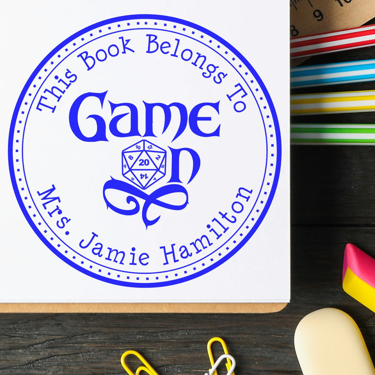 Blue custom book-ownership stamp seal with circular border, dice icon, and names on white paper beside colorful stationery.