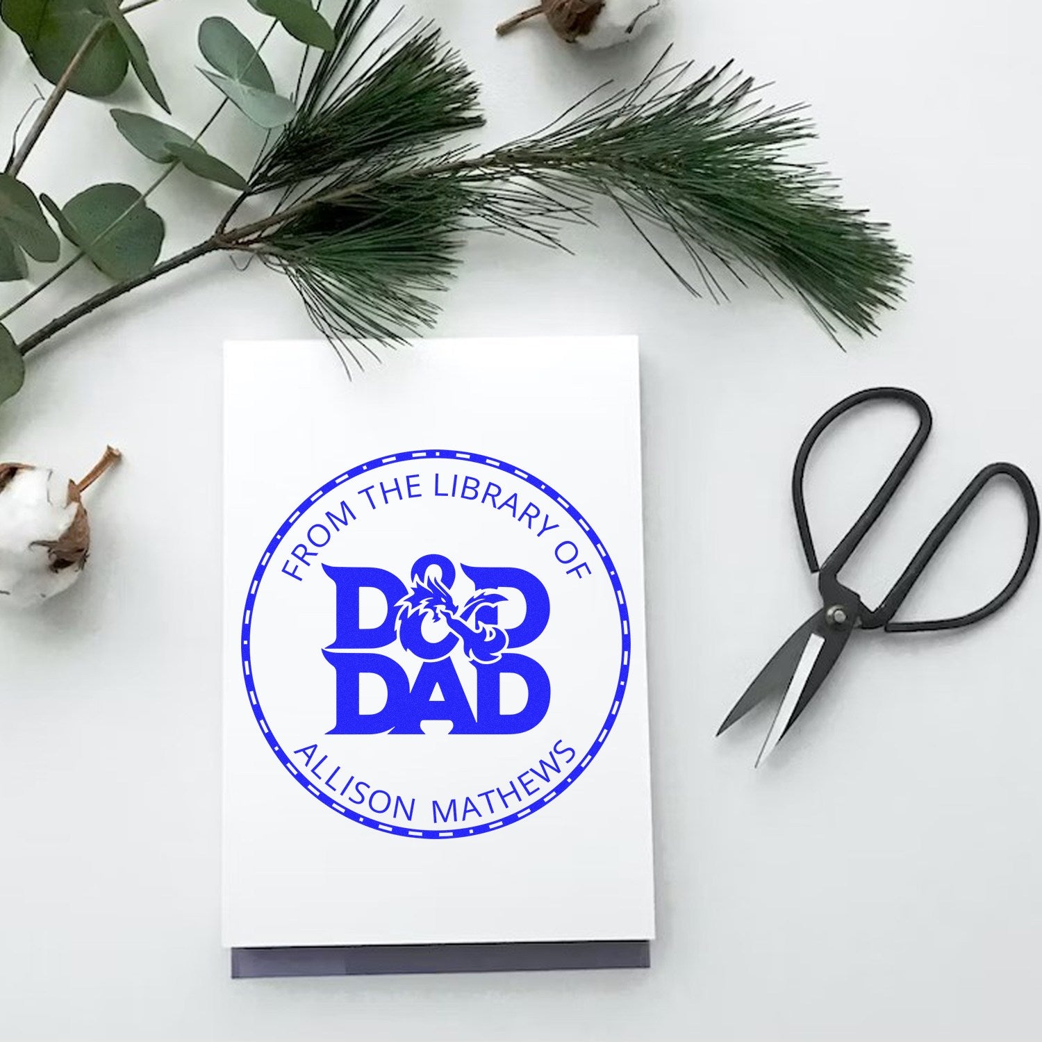 Blue personalized rubber stamp seal on white card, with pine branches and black scissors on a white background.