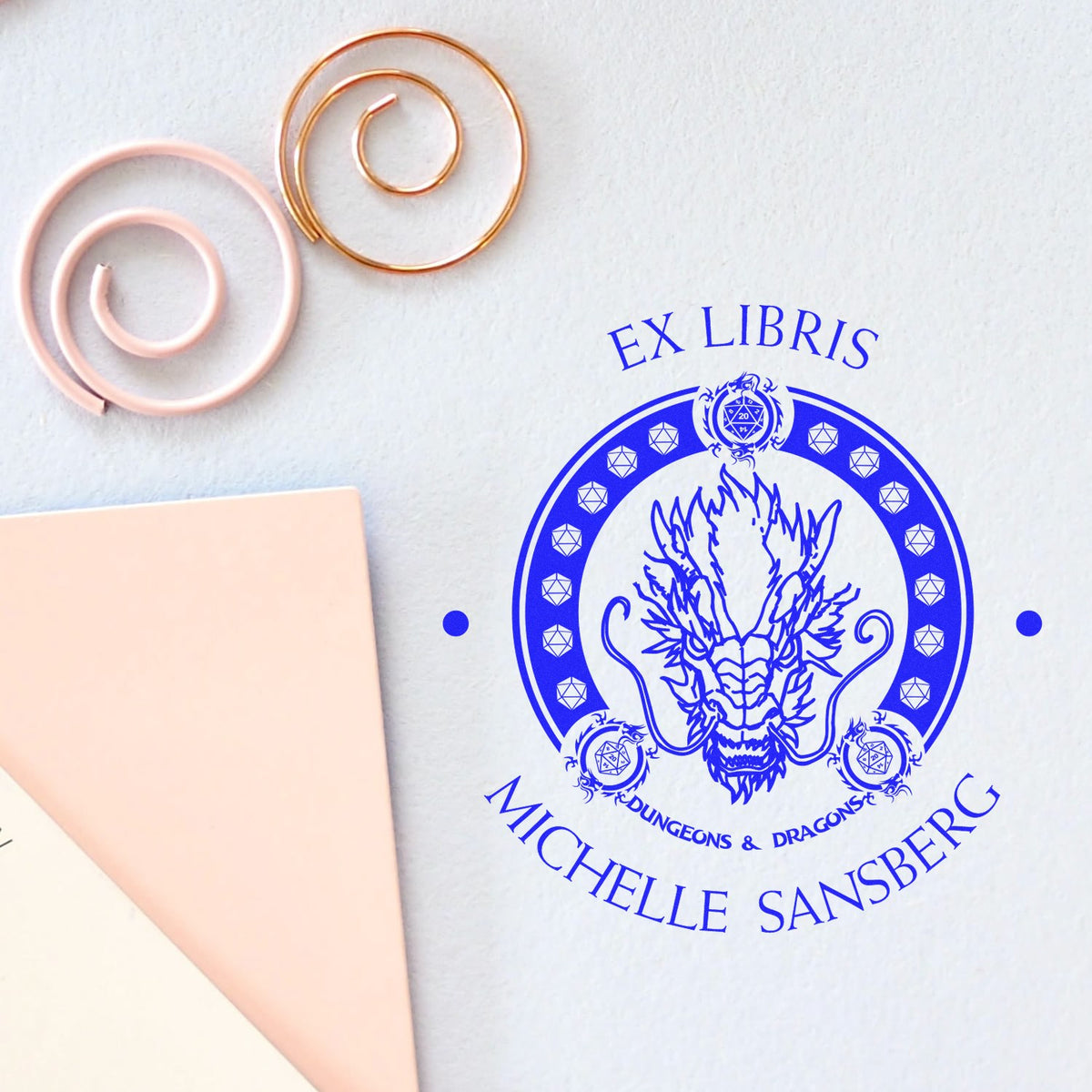 Blue ex libris stamp seal with dragon crest, plus blush pink and copper wax seal spirals on a light background.