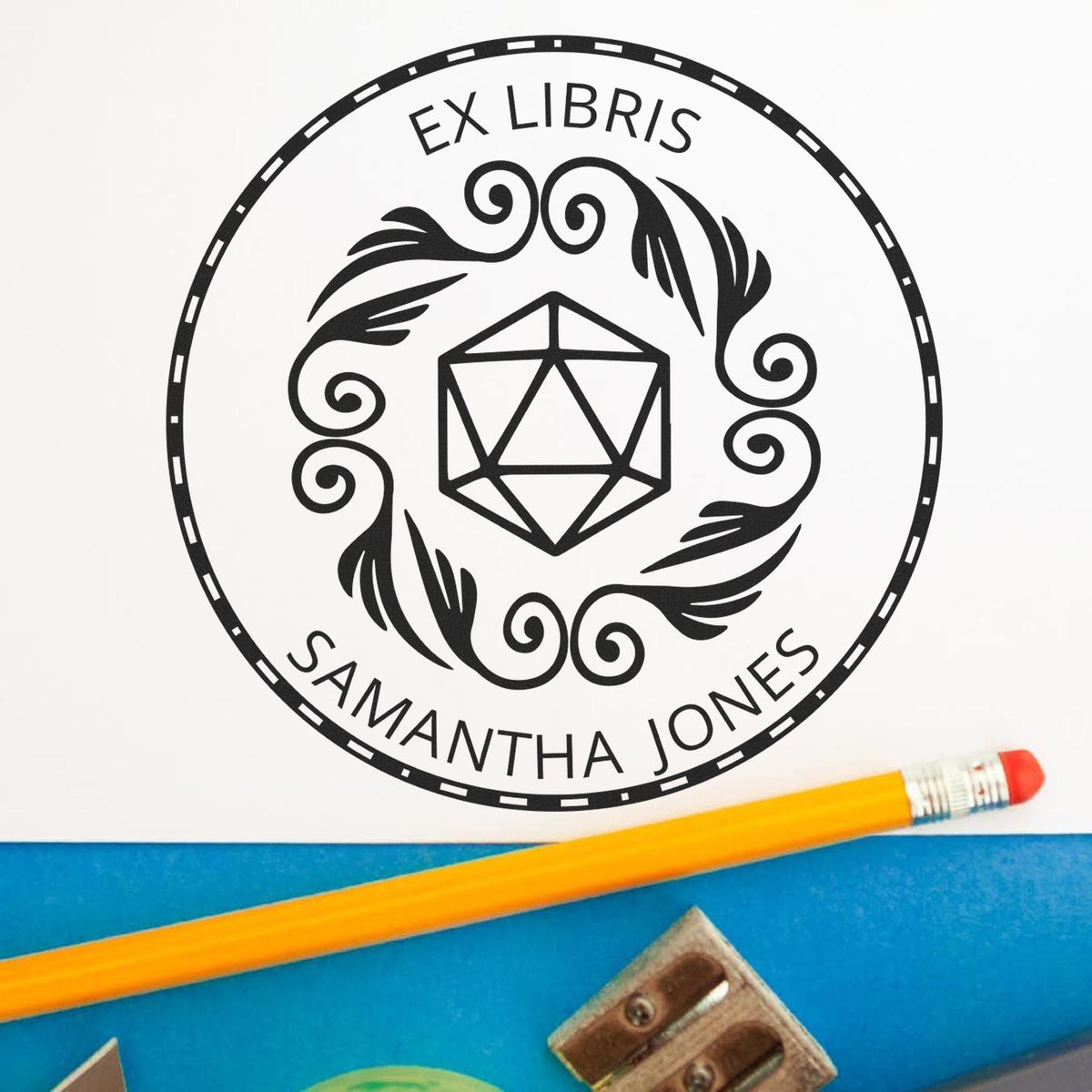 Black Ex Libris stamp seal with Samantha Jones, geometric icon, on white paper beside a yellow pencil and blue desk.