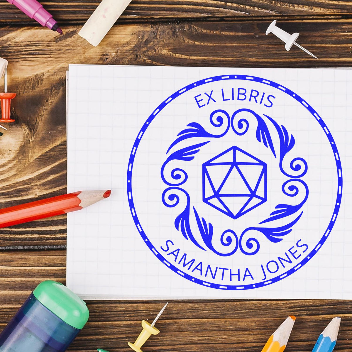 Blue custom ex libris stamp seal with geometric emblem and Samantha Jones on white paper, surrounded by pencils on rustic wood.