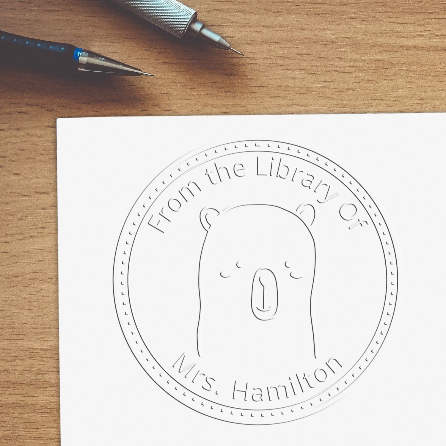 A Soft WhiskerTwist cartoon animal embossing seal is pressed onto paper, featuring a bear design with the text "From the Library Of Mrs. Hamilton." Nearby are pens and a customizable address stamp, part of a custom-made book collection.