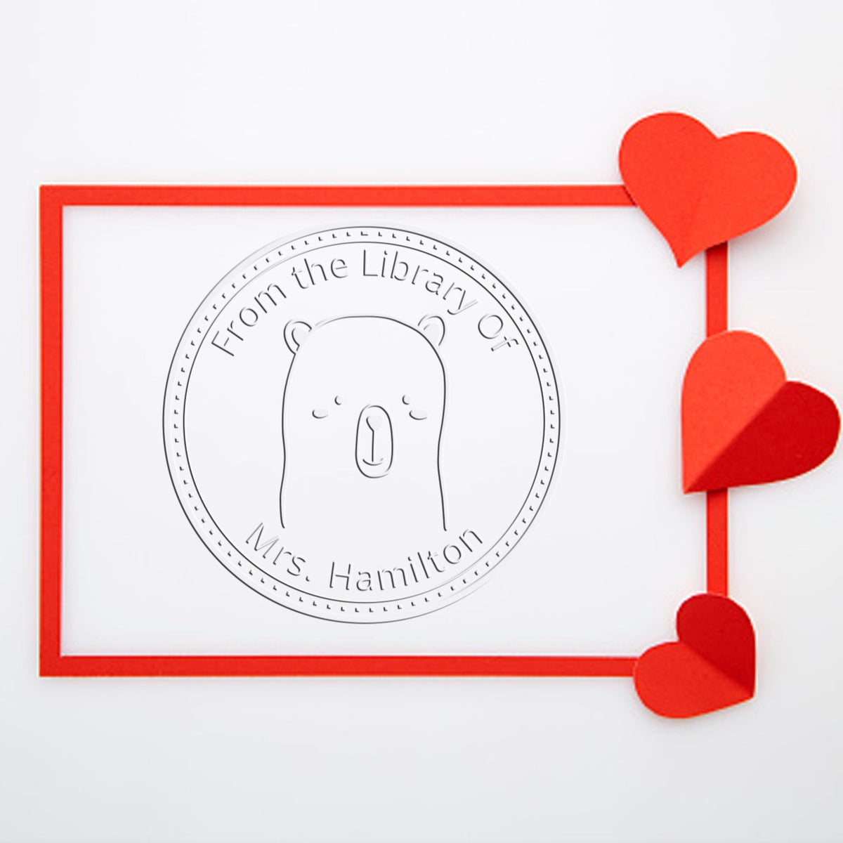 WhiskerTwist cartoon animal embosser on a desk, featuring a custom-made library seal with a bear design. Includes a personalizable return mail stamp, framed by a red border with heart accents.