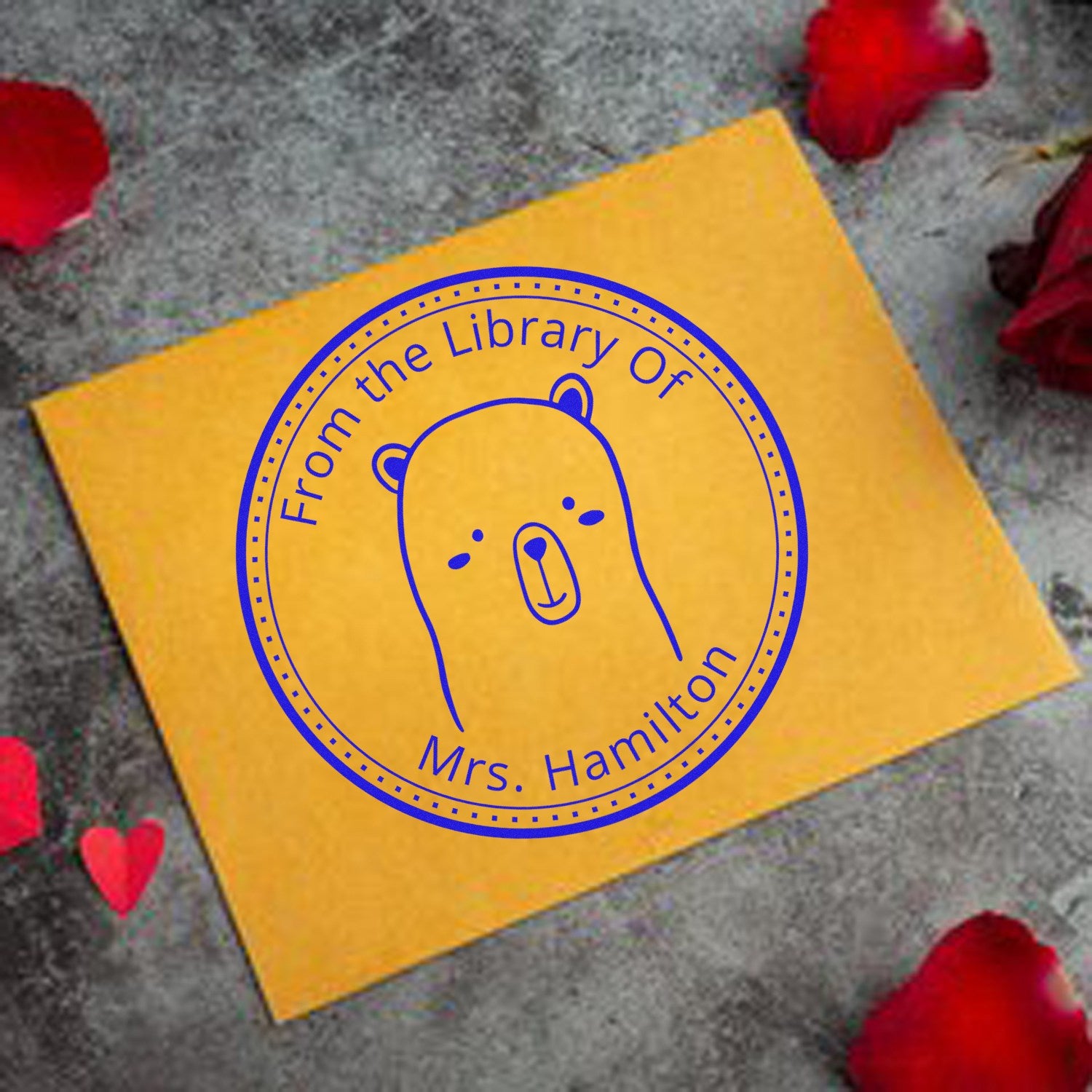 Yellow envelope with a blue stamp featuring a cartoon animal with a wood handle whisker twist design. The stamp reads "From the Library Of Mrs. Hamilton." Surrounded by red rose petals, it's a personalized book lover gift and custom-made home stamp.
