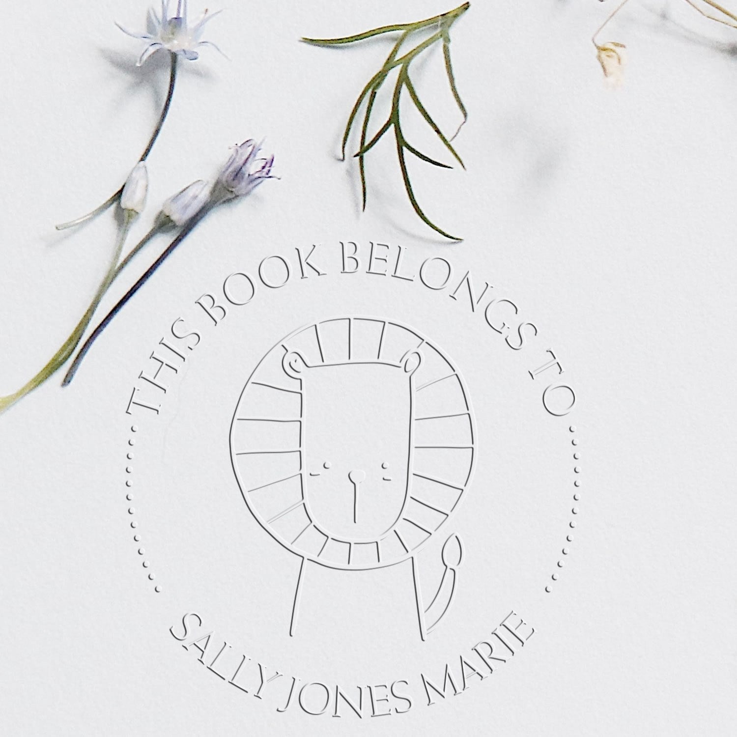 Soft ZippyZap cartoon animal custom-made book embossing seal on paper, featuring a lion design with text "This Book Belongs To Sally Jones Marie." Surrounded by delicate flowers, showcasing a personalized home stamper.
