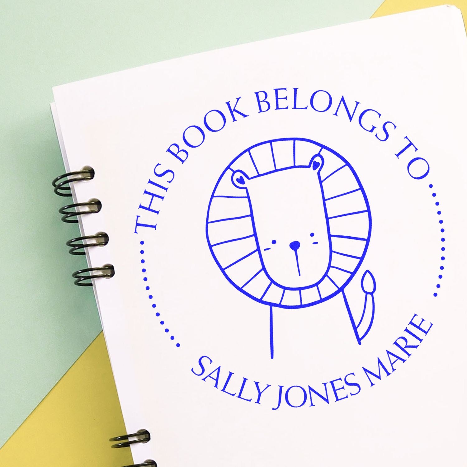 Image of a spiral notebook with a blue cartoon animal stamp featuring a lion and the text "This Book Belongs To Sally Jones Marie." The stamp has a wood handle. Keywords: Wood Handle ZippyZap Cartoon Animal Personalized Book Belongs To Stamp, Personalizable New Address Stamp.