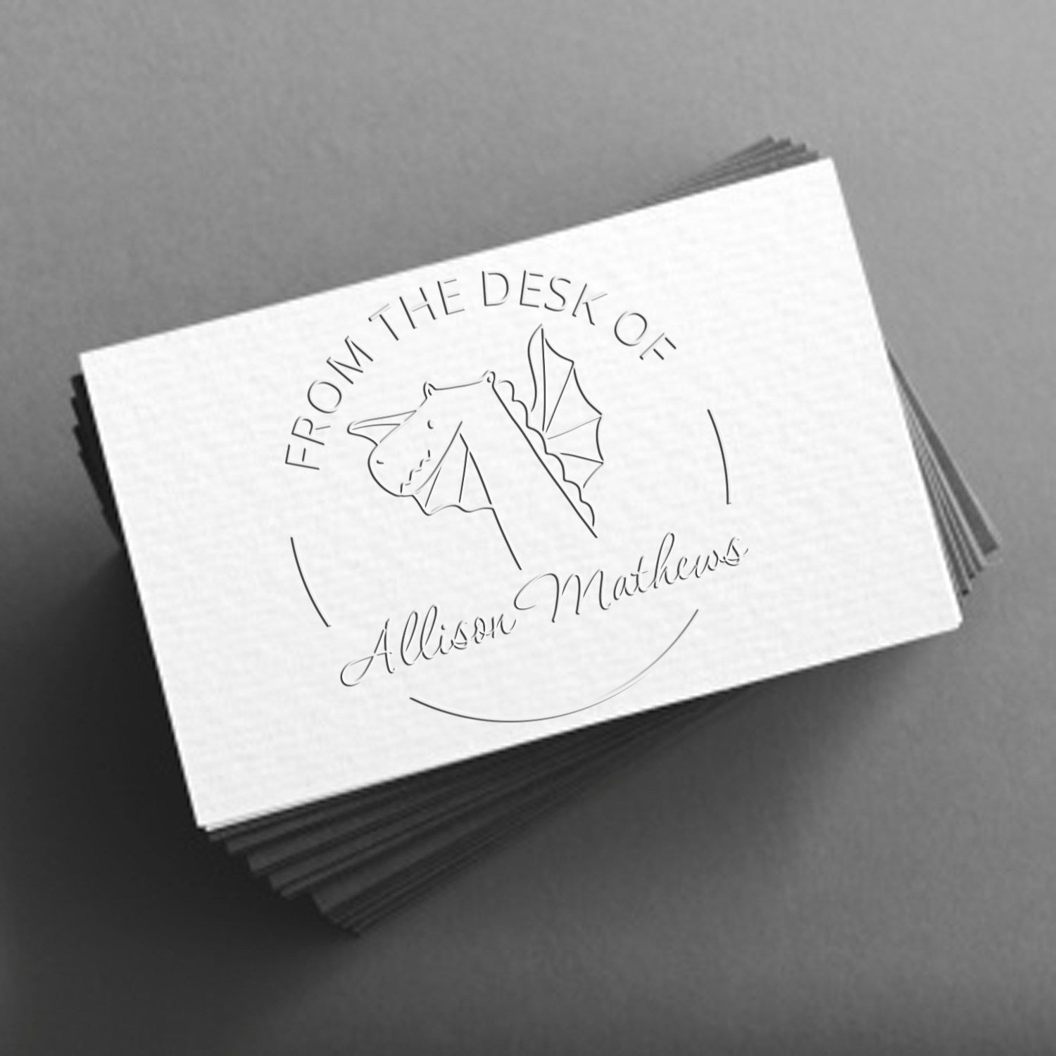 A stack of white cards embossed with a Soft SnuggleSnout cartoon animal design, featuring a dragon and the text "From the Desk of Allison Mathews." The image showcases a custom-made book marking embosser and customizable return mail stamper.