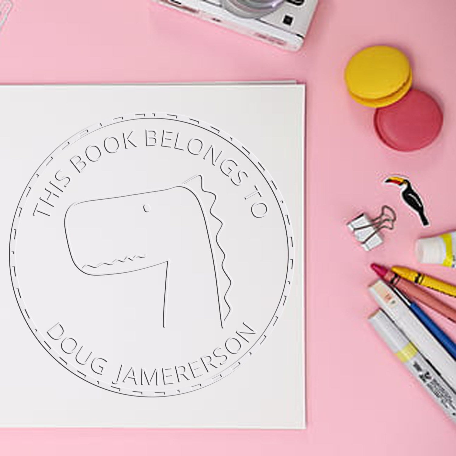 A Soft JellyBeanJump cartoon animal embossing seal is pressed onto a book page, reading "This Book Belongs To Doug Jamerson." Nearby are colorful pens and a Custom-Made Address Stamp on a pink surface.