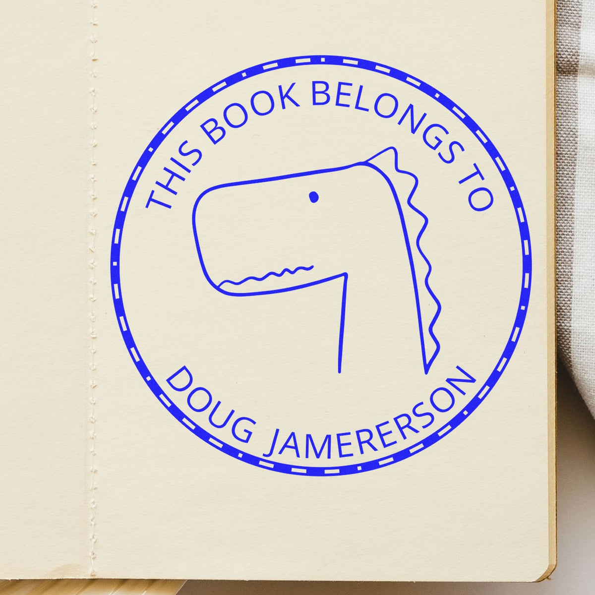 Slim Pre-Inked JellyBeanJump Cartoon Animal Custom Bookplate Design Stamp featuring a simple dinosaur illustration and the text "This Book Belongs To Doug Jamerson" in blue. Custom Stamp is applied on an open book page.