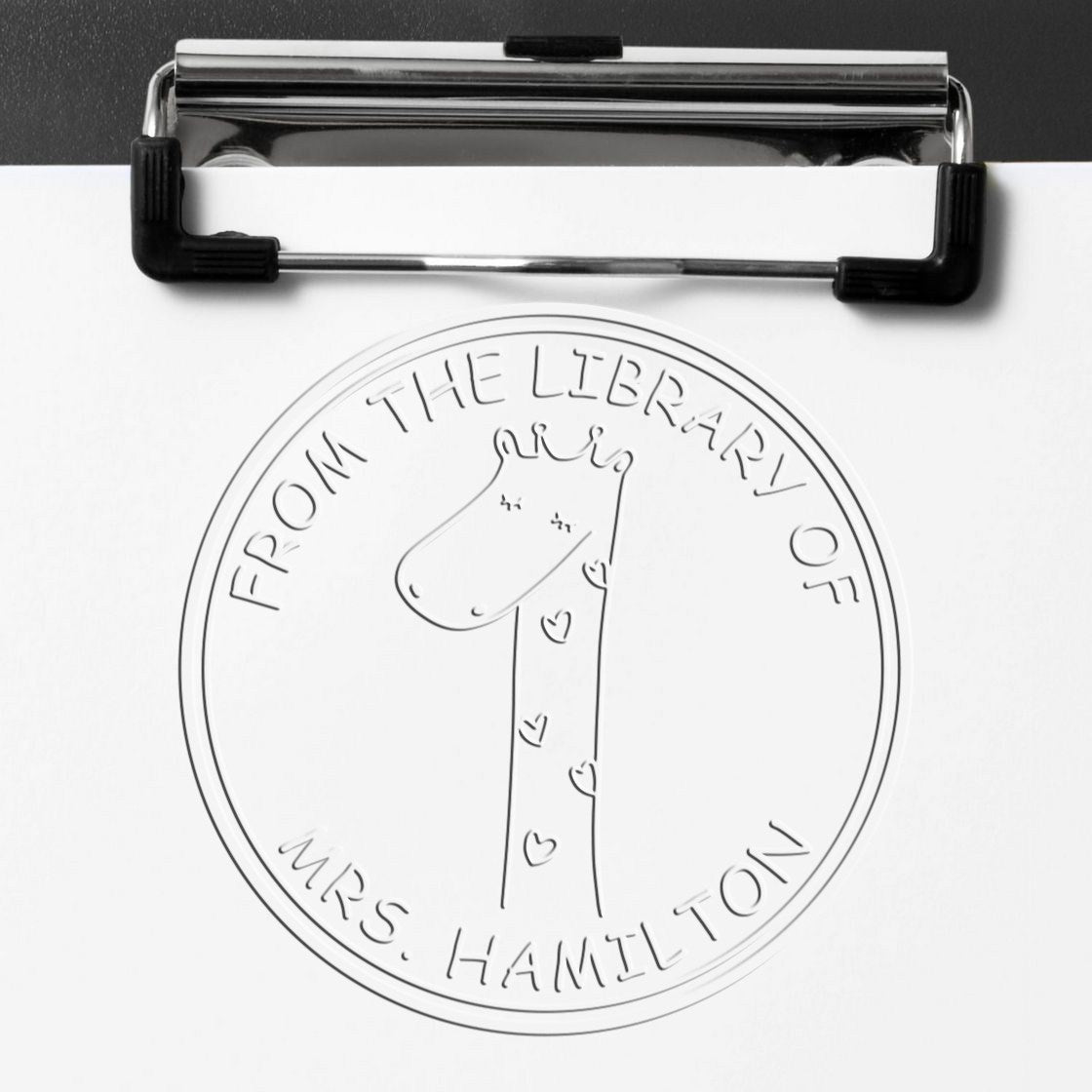 A custom-made book signature embosser featuring a cartoon animal with a soft squiggle design, resembling a giraffe with a crown and heart patterns. The text reads From the Library of Mrs. Hamilton. A customized new address stamper is also visible.