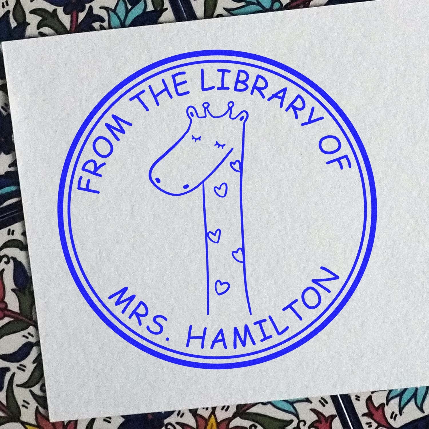 A Wood Handle SquiggleSquash Cartoon Animal Personalized Stamp featuring a cute giraffe with hearts, encircled by the text From the Library of Mrs. Hamilton. This Customizable Mailing Stamp is displayed on a patterned background.