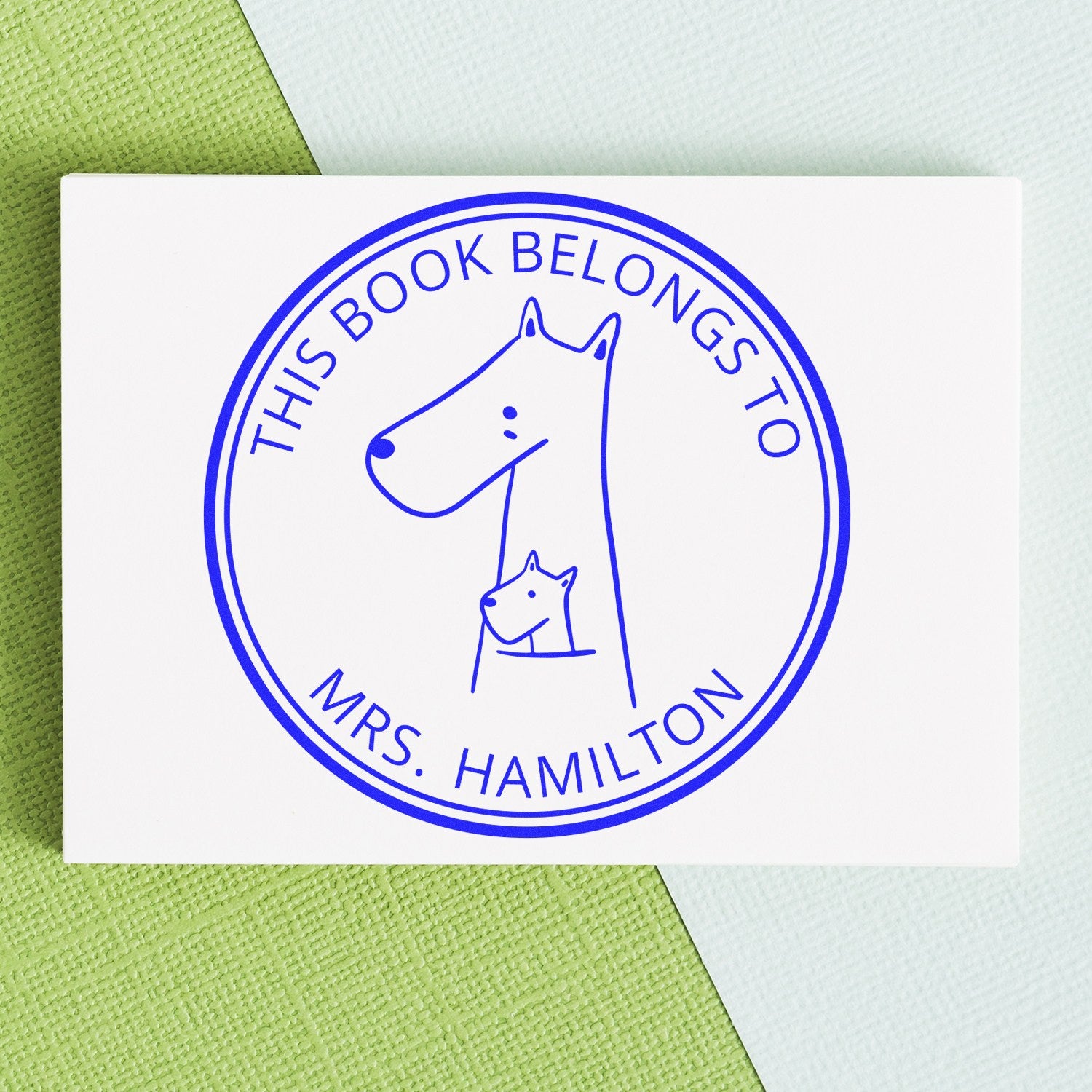 MuddleMoo Cartoon Animal Pre-Inked Customizable Book Plate Stamp featuring a simple animal design with text "This Book Belongs To Mrs. Hamilton." Personalized Home Stamper on a textured green and blue background.