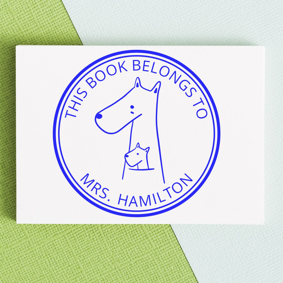 Slim Pre-Inked MuddleMoo Cartoon Animal Custom Bookplate Sticker Stamp featuring a simple animal design with text "This Book Belongs To Mrs. Hamilton." Ideal as a Custom Mailing Stamp, shown on a textured green and blue background.
