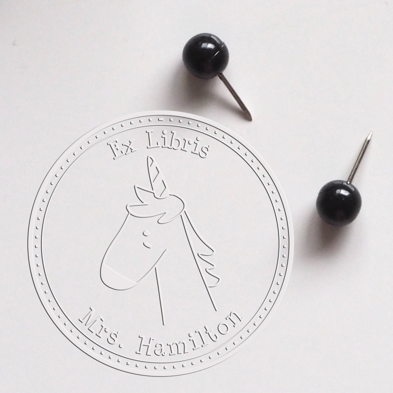 A Soft GlimmerGlide Cartoon Animal Custom-Made Book Signature Embossing Seal featuring a unicorn design with "Ex Libris" and "Mrs. Hamilton" text. Nearby are two black pushpins. Also shown is a Custom-Made New Address Stamper.