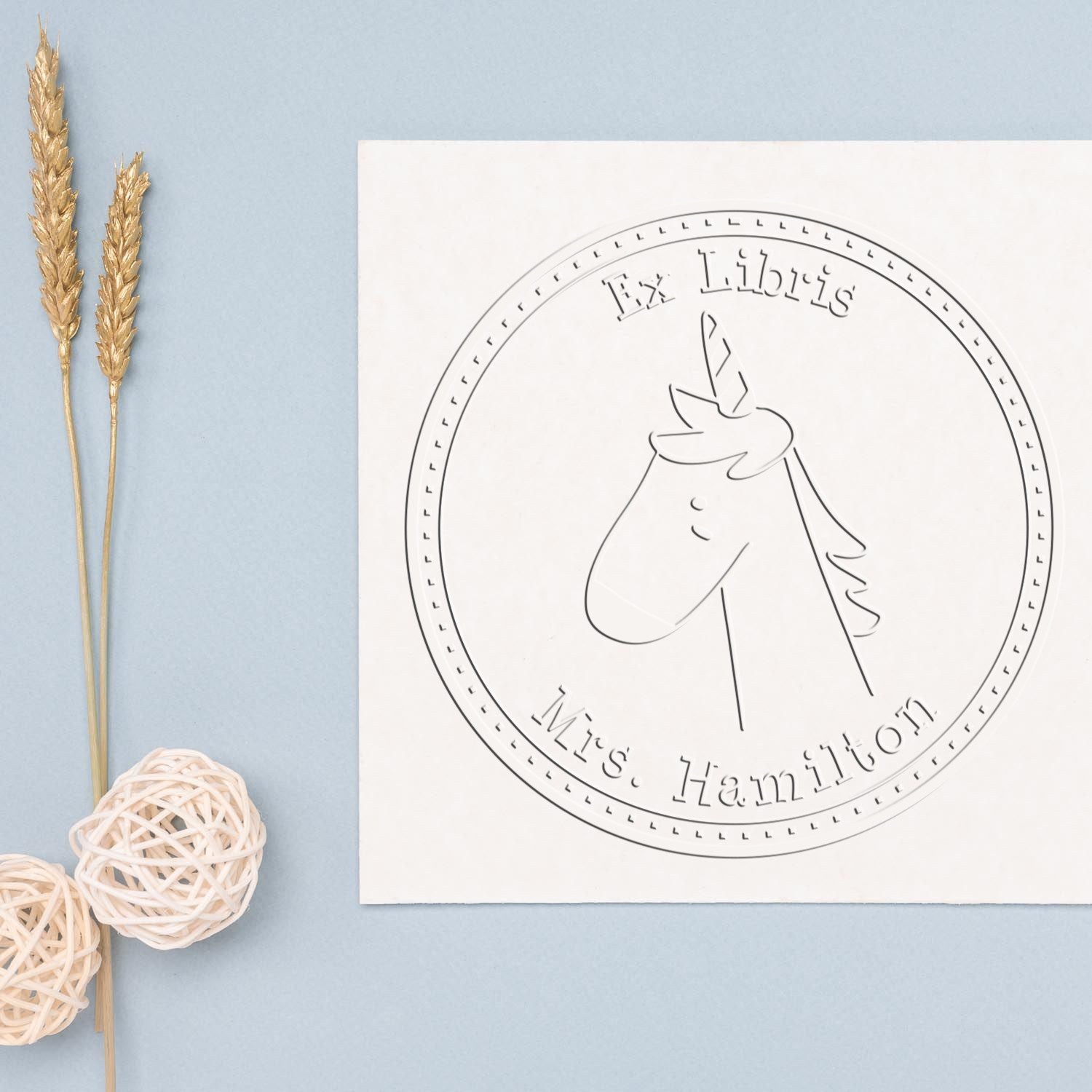 A GlimmerGlide Cartoon Animal Handheld Personalized Ex Libris Book Embosser imprint on paper, featuring a unicorn design with "Ex Libris" and "Mrs. Hamilton" text. Nearby are decorative elements, showcasing a Customized Home Stamper.