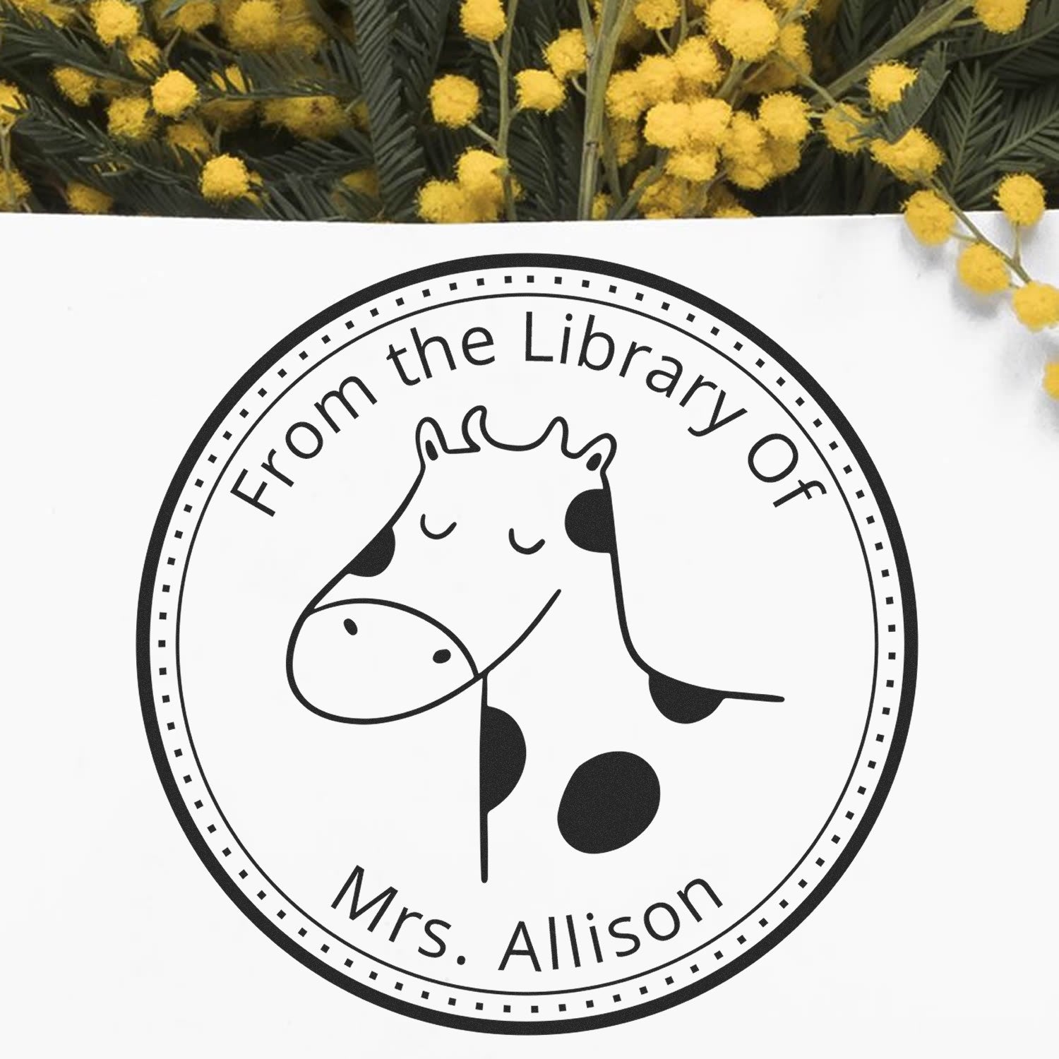 Cartoon animal stamp with a smiling giraffe and text "From the Library Of Mrs. Allison." The BoopSnoot self-inking, custom-made stamp is used on paper, surrounded by yellow flowers. Keywords: Customized Home Stamp, Belongs To The Library Of.