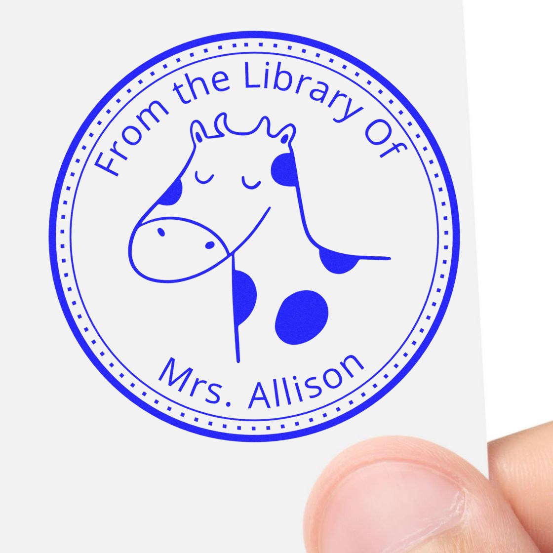 A hand holds a white card stamped with a blue Slim Pre-Inked BoopSnoot Cartoon Animal Custom Bookplate Printing Stamp featuring a smiling cow. The text reads "From the Library Of Mrs. Allison." Ideal for a Customizable Address Stamp.